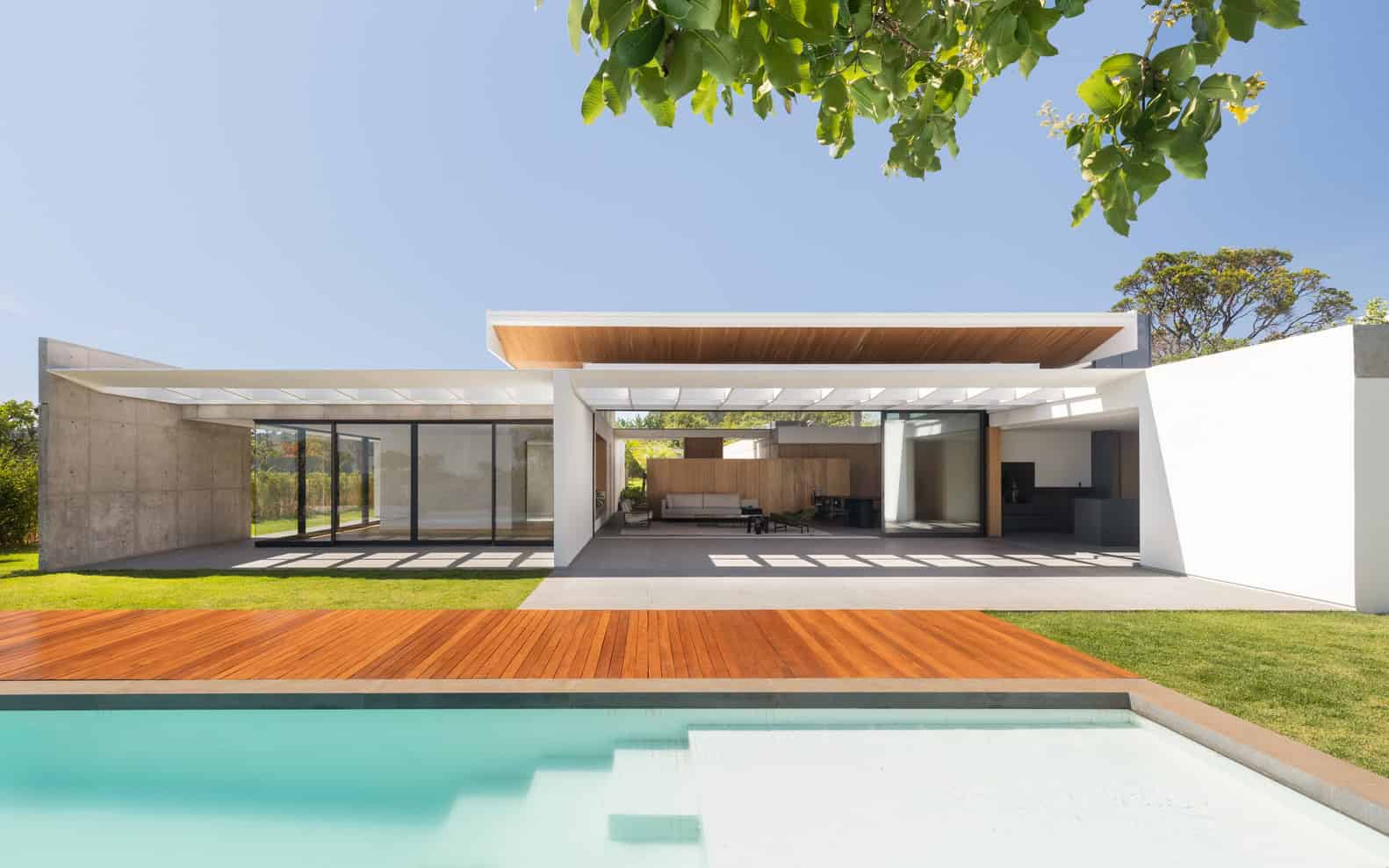 Modern minimalist house with open-concept living space, large glass windows, and seamless indoor-outdoor design, set against a lush green landscape with a swimming pool.