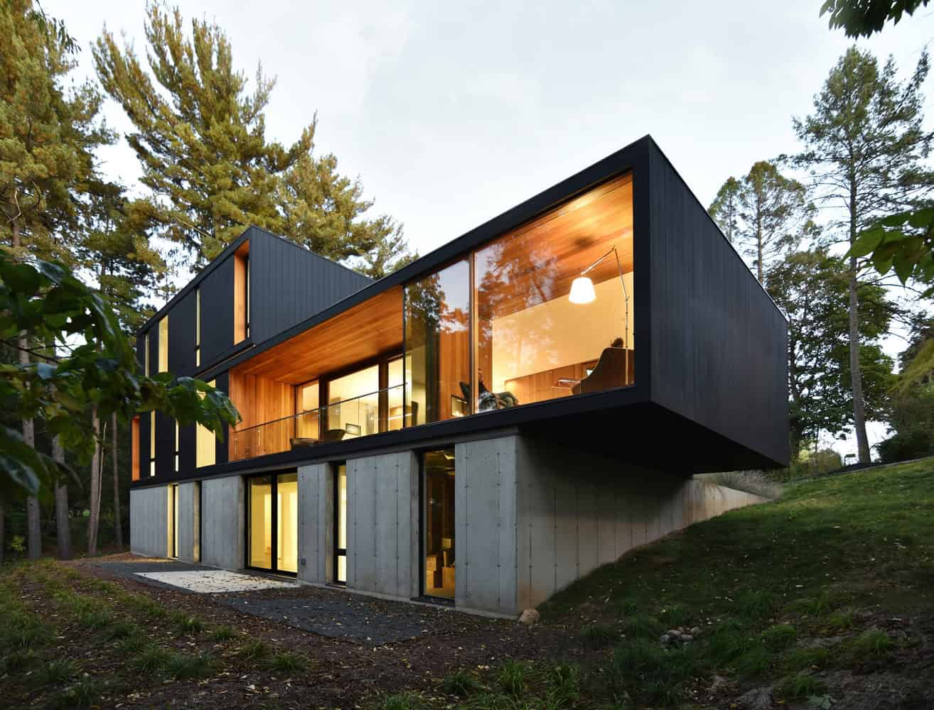 Contemporary modern house with large glass windows, timber accents, and a concrete base nestled in a lush, green forest setting.