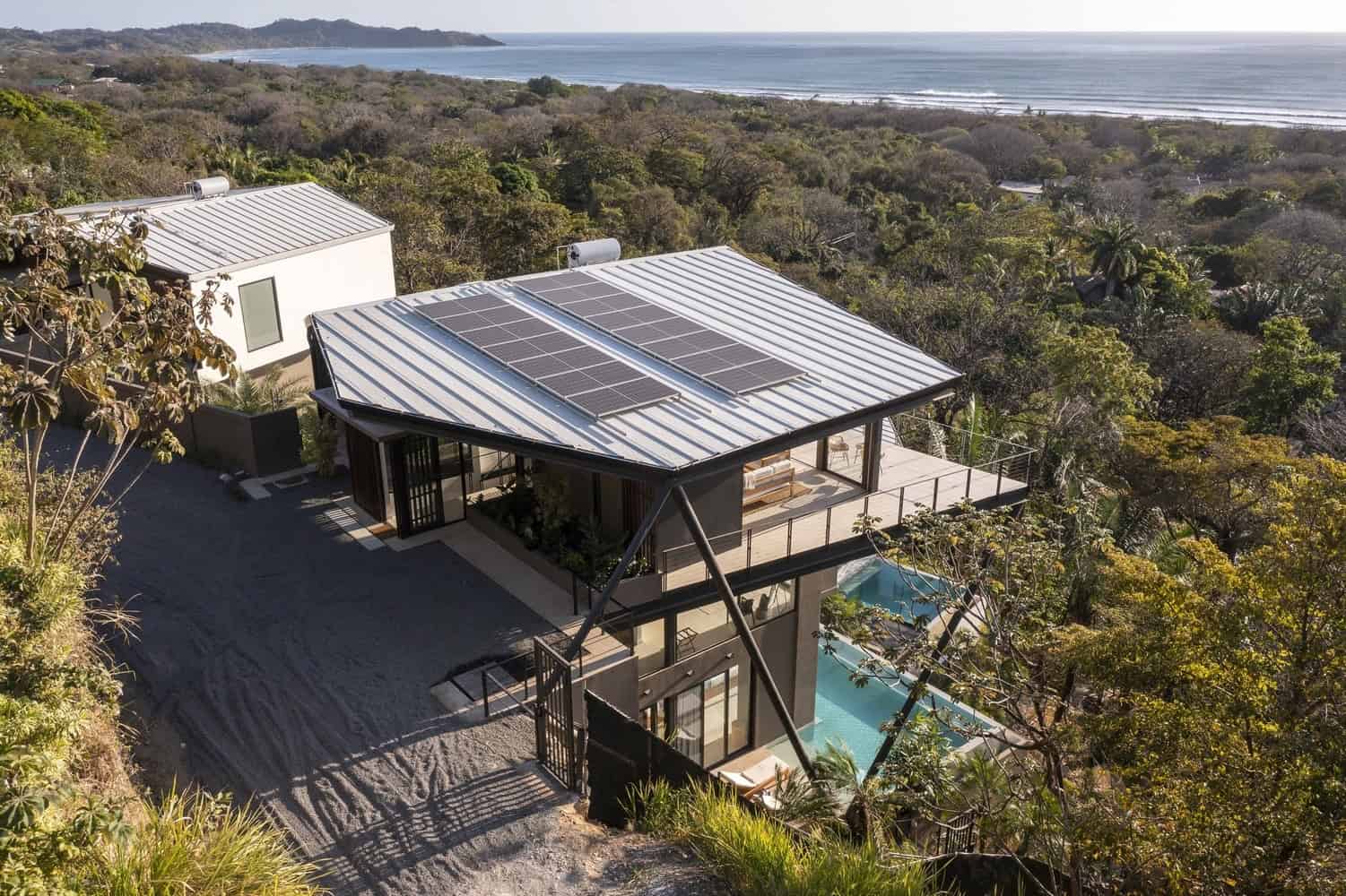 Modern hillside home with solar panels on metal roof, surrounded by lush greenery and ocean views, exemplifying innovative eco-friendly architecture and sustainable design.