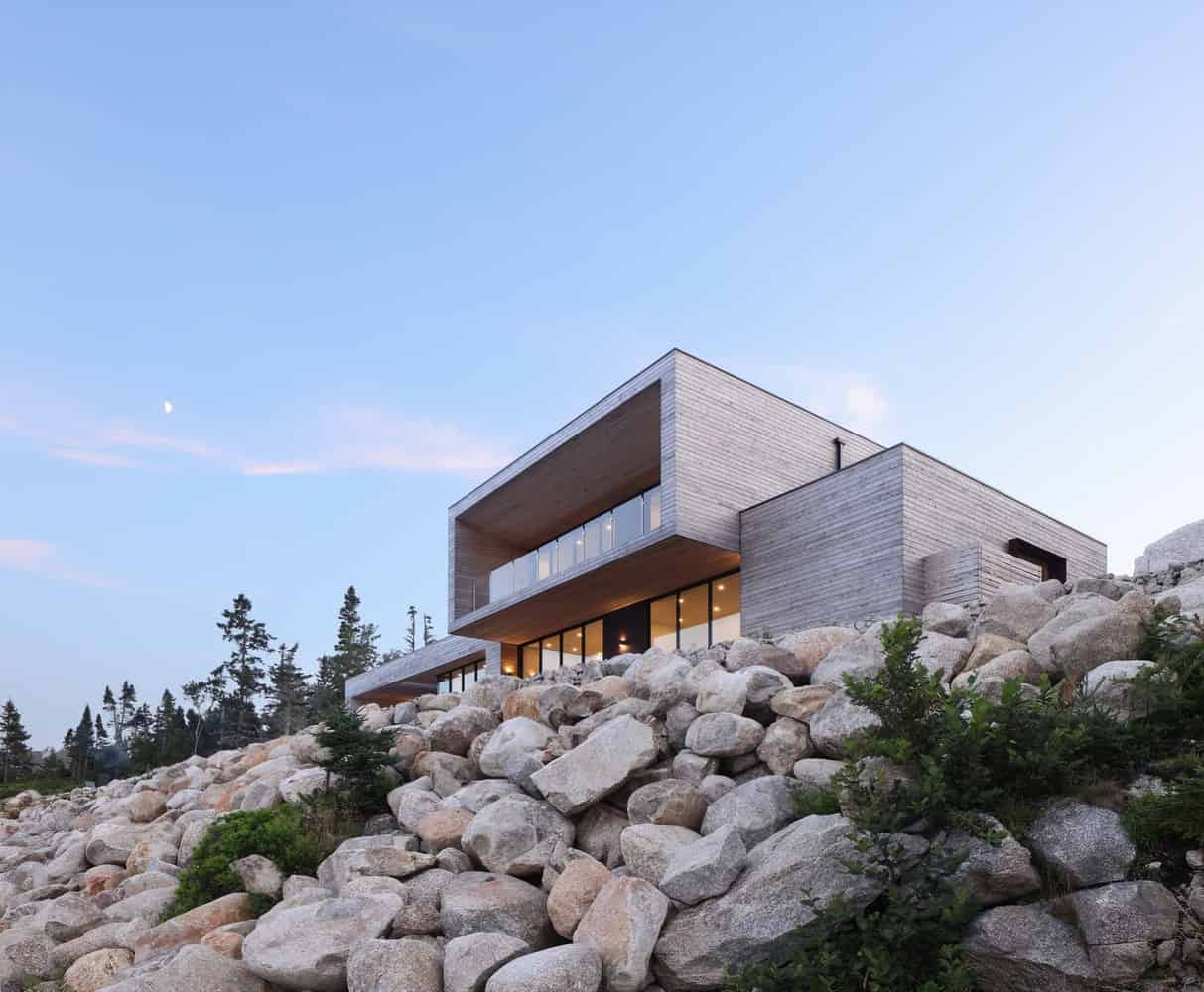 Modern minimalist house built on rocky terrain with large glass windows and concrete exterior, situated in a natural landscape with trees and a clear sky.