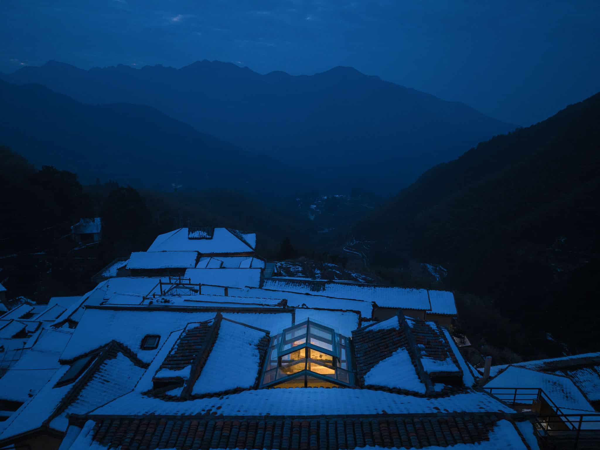 Snow-covered rooftops with mountain landscape in the background during dusk, showcasing rustic architecture and serene winter scenery.