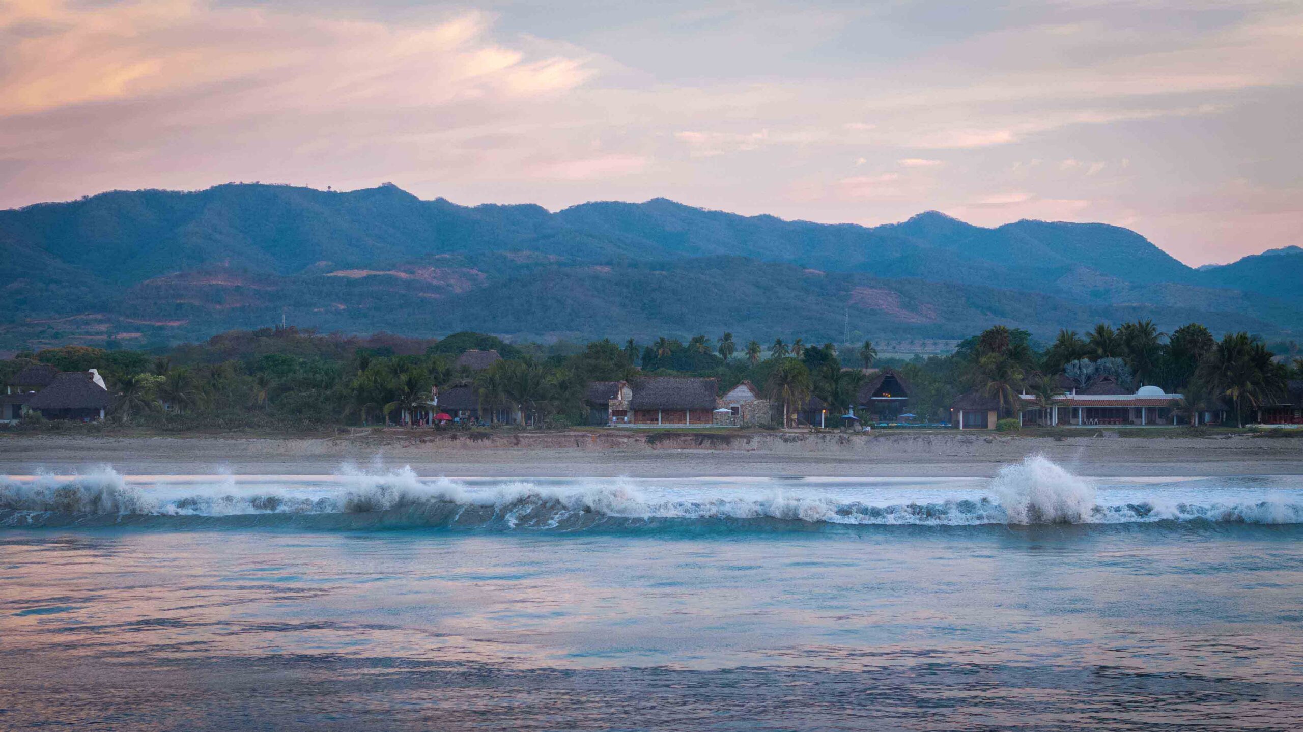 Tate House / MATERIA / Mexico Serene coastal landscape with tropical beachfront villas, lush greenery, and majestic mountains in the background, showcasing tranquil ocean waves at sunset, perfect for tropical architecture and vacation home designs.