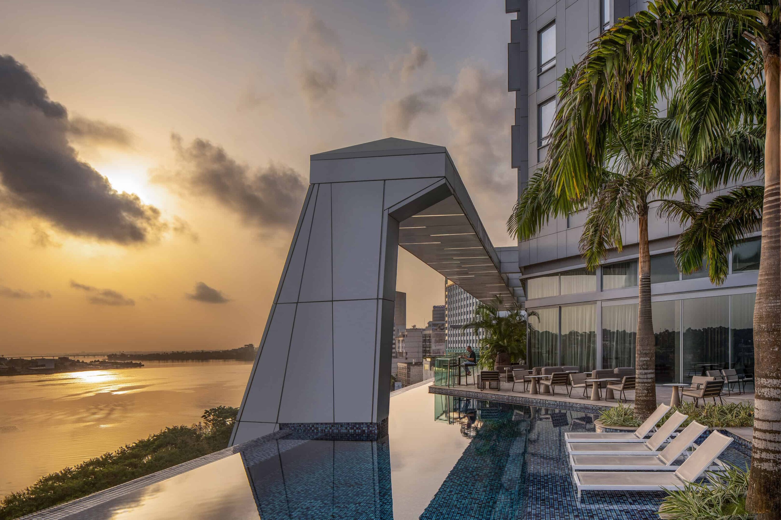 Luxury modern hotel rooftop with infinity pool overlooking city skyline during sunset, featuring contemporary architecture, tropical palm trees, and outdoor lounge area, ideal for architecture and design photography.