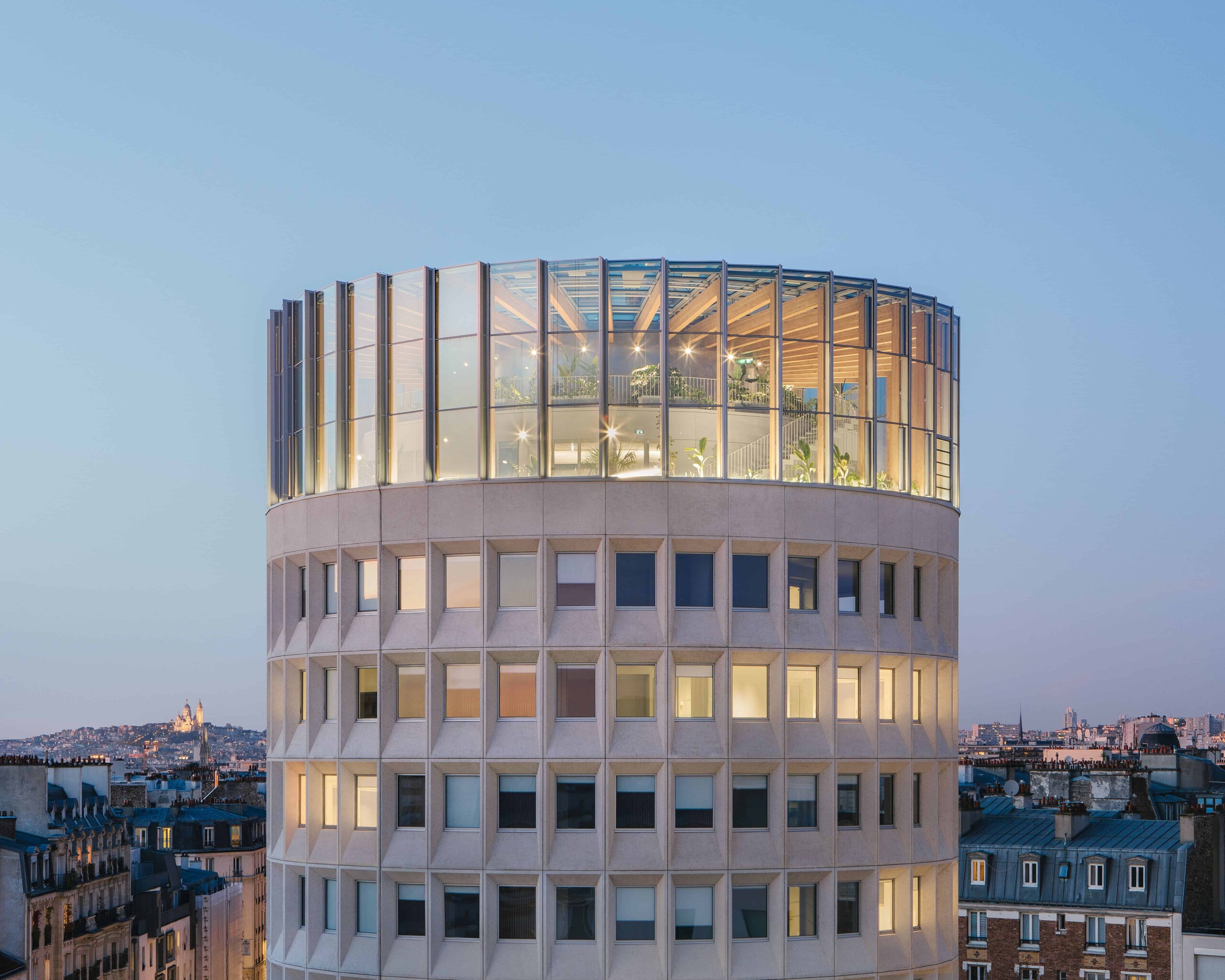 Modern circular glass office building with illuminated interior, set against a cityscape at dusk. Features contemporary architecture with large glass windows and a rooftop garden.