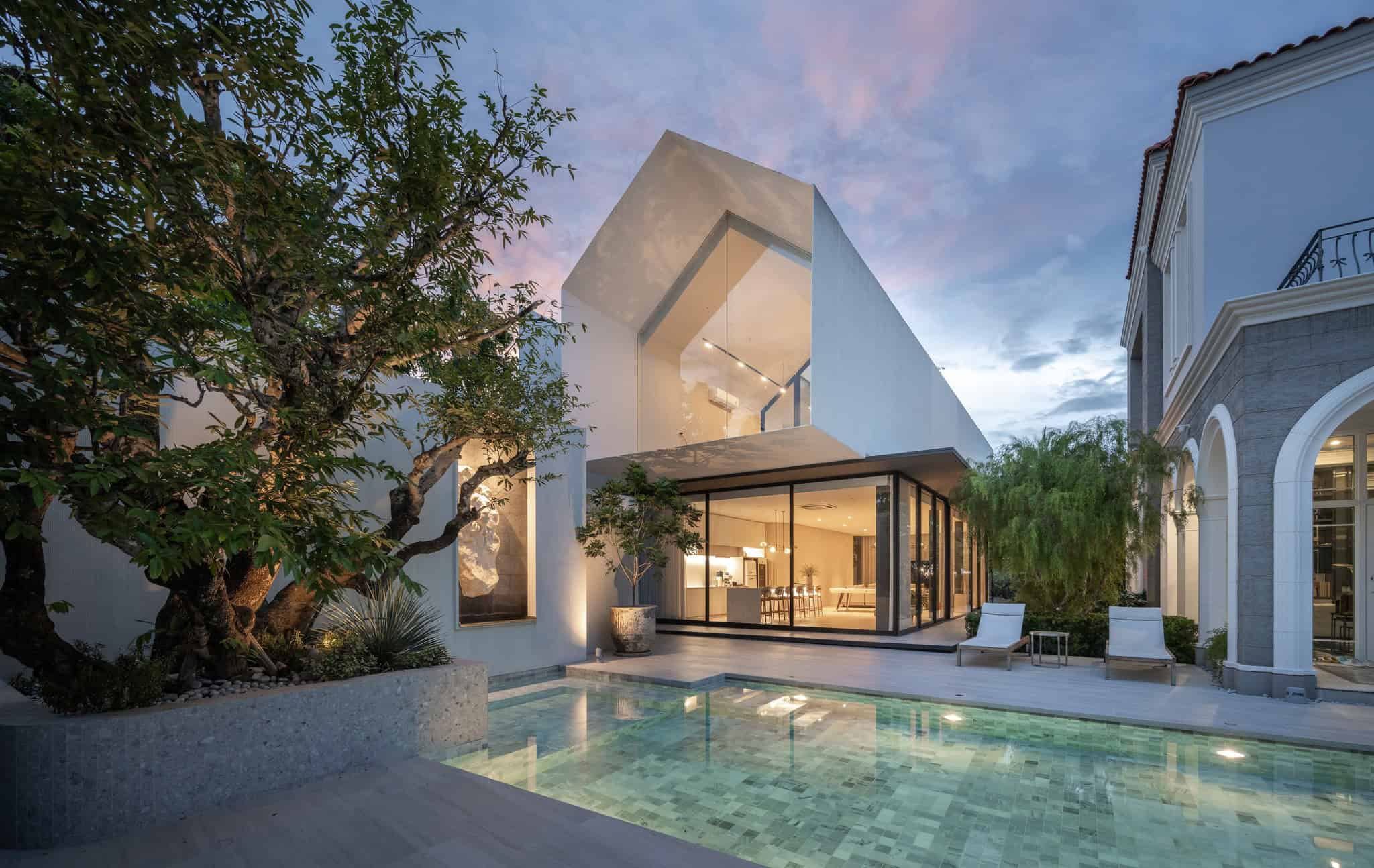 Contemporary modern house with sleek architecture, large glass windows, and a private pool surrounded by lush greenery at dusk.