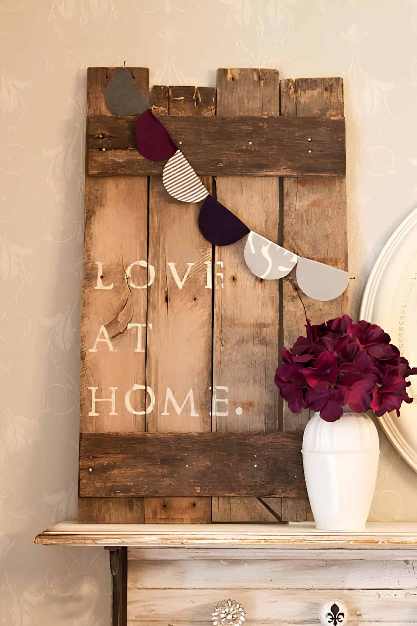 Vintage wood plank wall art with the phrase 'Love at Home' beside a vase of deep red flowers.