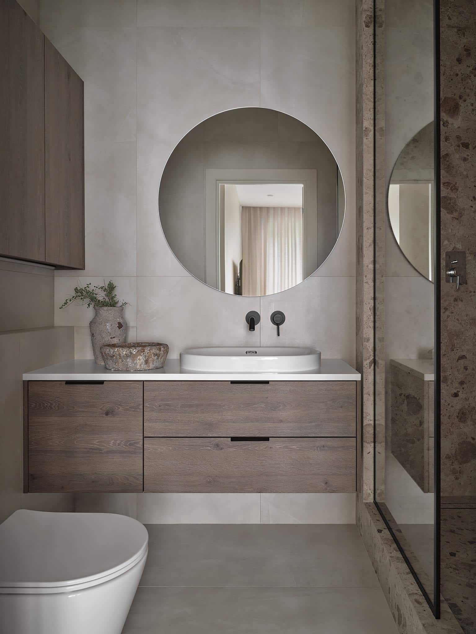 Modern Bathroom with Round Mirror and Wood Vanity Modern bathroom with round mirror, stone wall tiles, and wood vanity under basin sink.