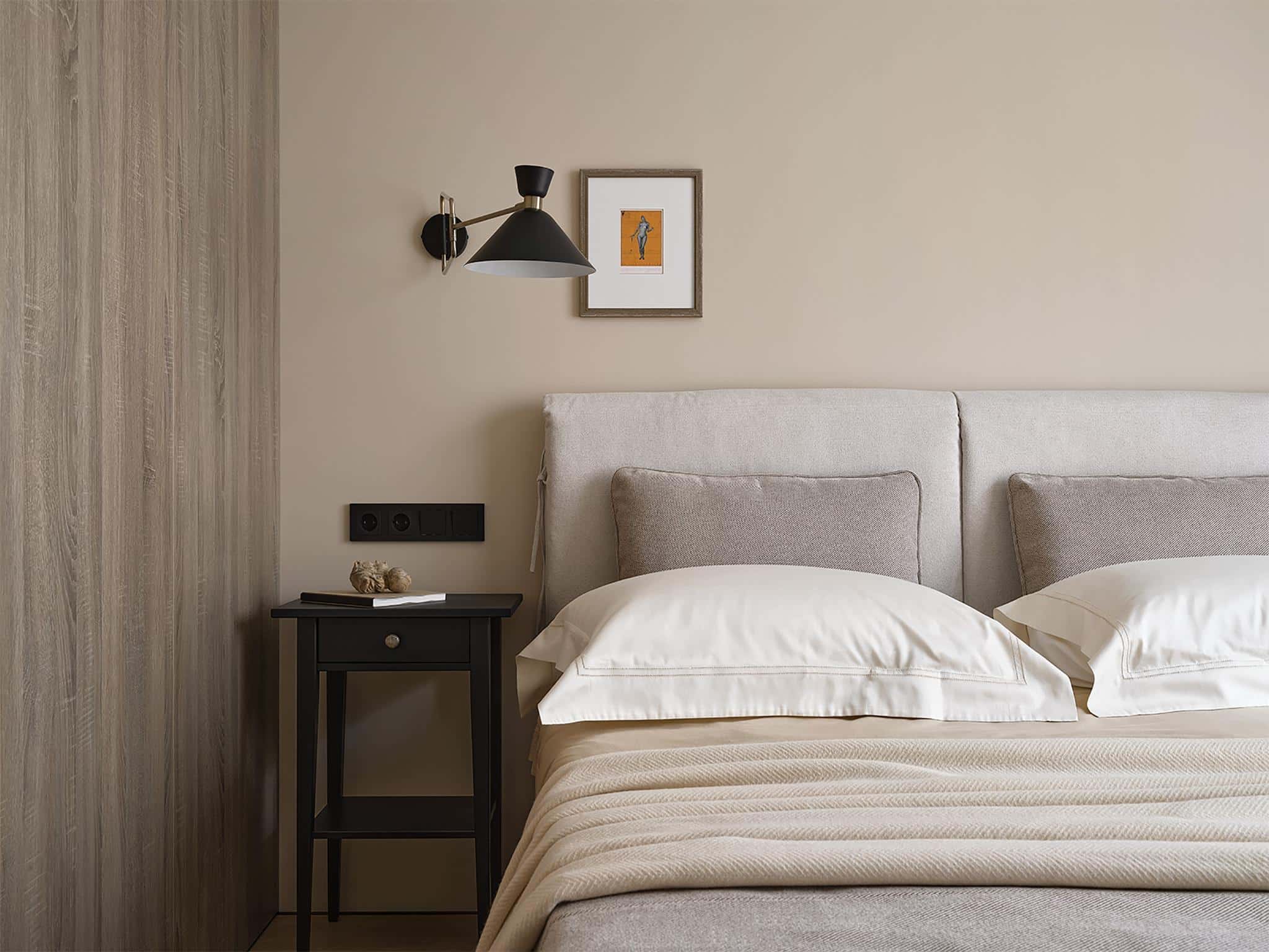 Neutral Bedroom Wall Lamp and Art Detail Neutral bedroom with fabric headboard, black wall lamp, and framed artwork above nightstand.