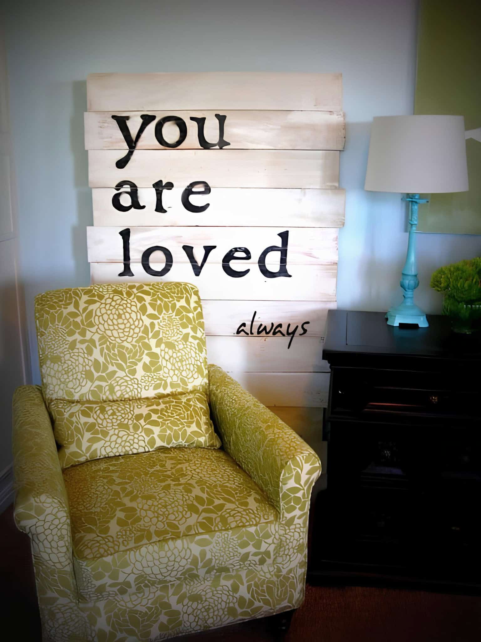 Whitewashed wood wall sign with 'you are loved always' quote beside an upholstered green floral armchair.