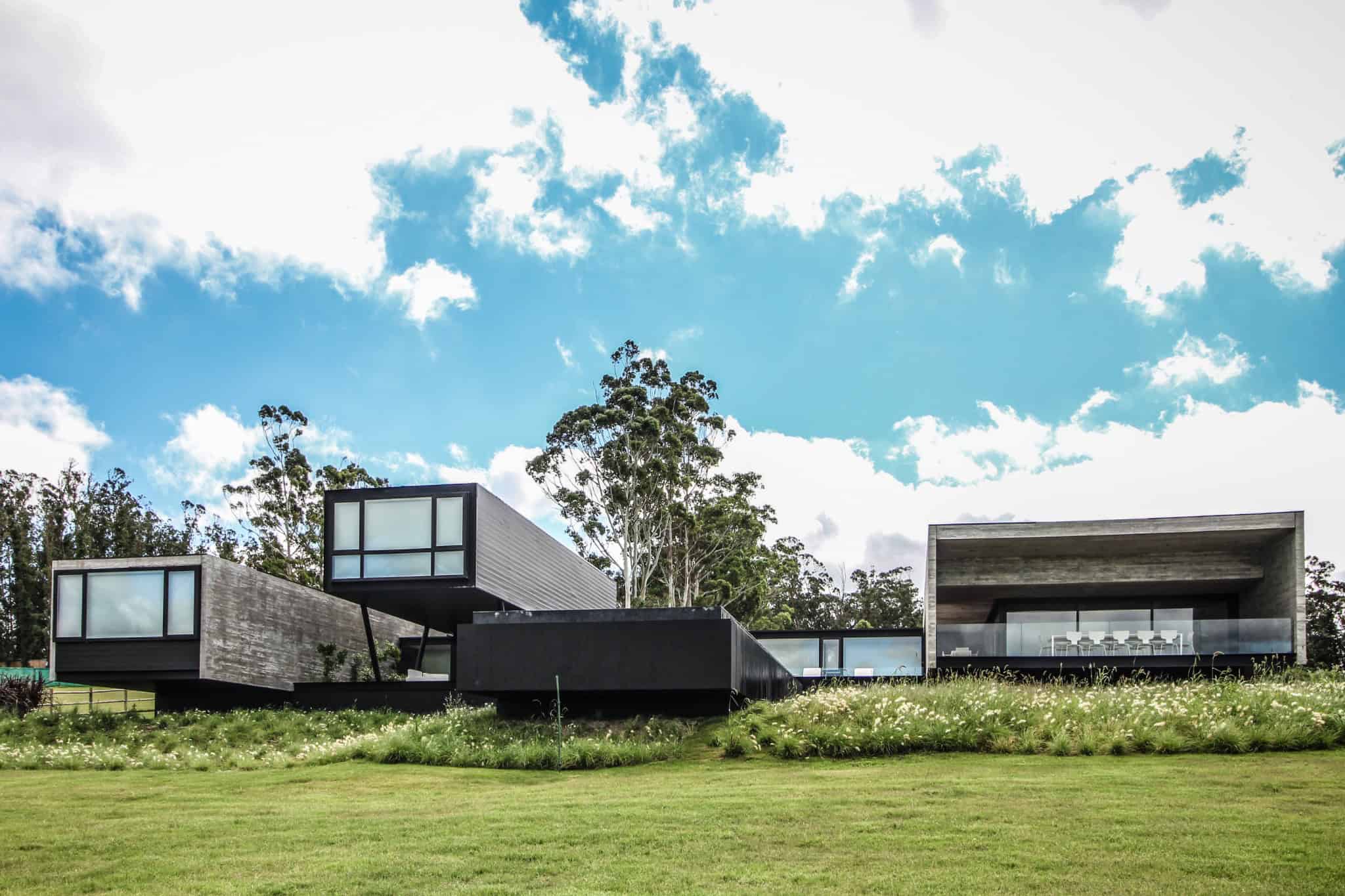 Contemporary modern house with geometric design, large glass windows, and concrete elements set against a lush green landscape and partly cloudy sky.