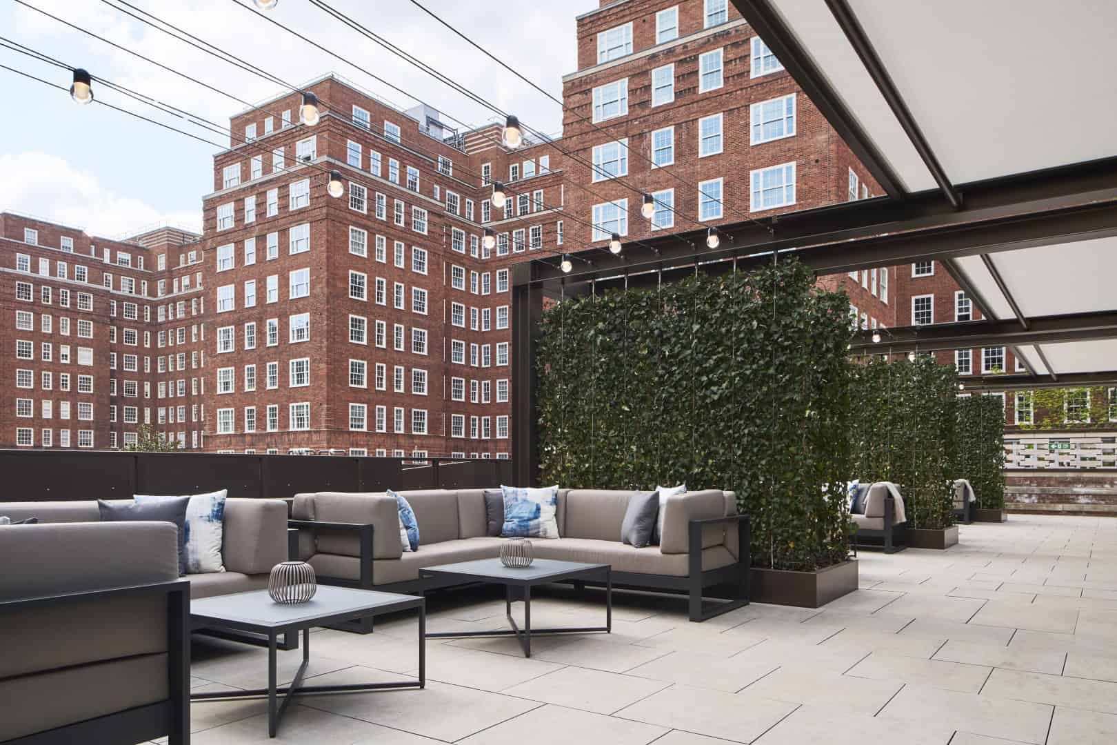 Modern outdoor rooftop terrace with contemporary furniture, string lights, and cityscape view featuring a red brick building, ideal for urban outdoor living and stylish relaxation.