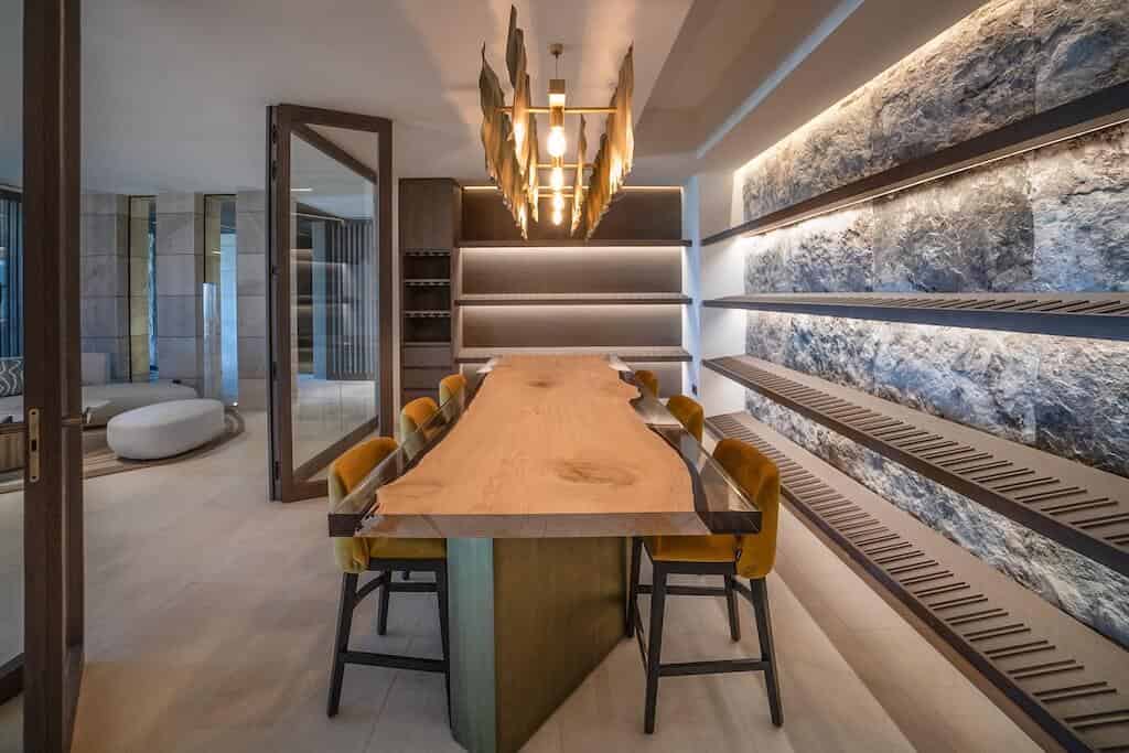 Villa Kaizen Live Edge Wine Table Wine cellar in Villa Kaizen with live-edge table, backlit stone wall, and open shelving.
