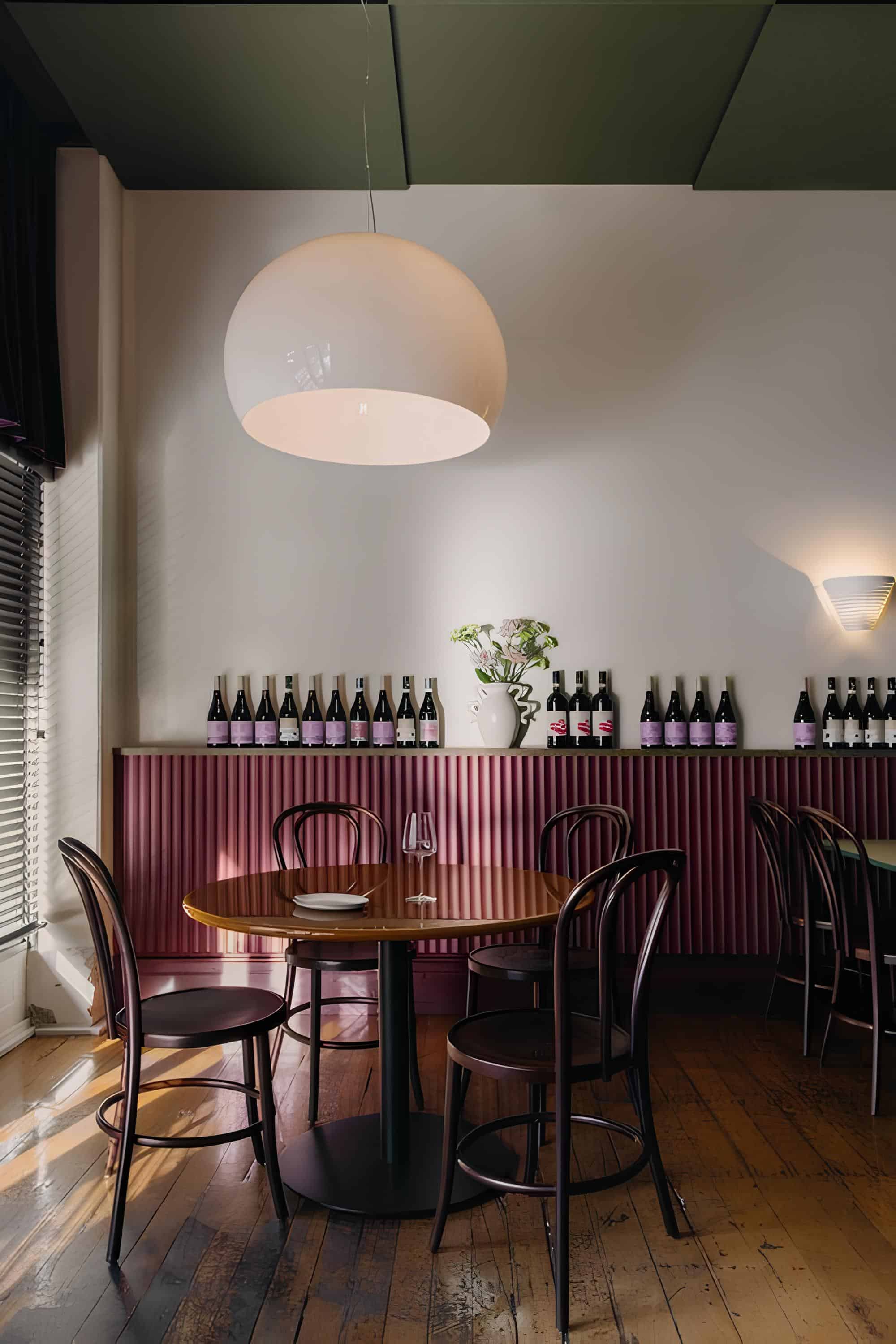 Round table under large pendant light at Vin Populi Next Door, with wine bottles along wall