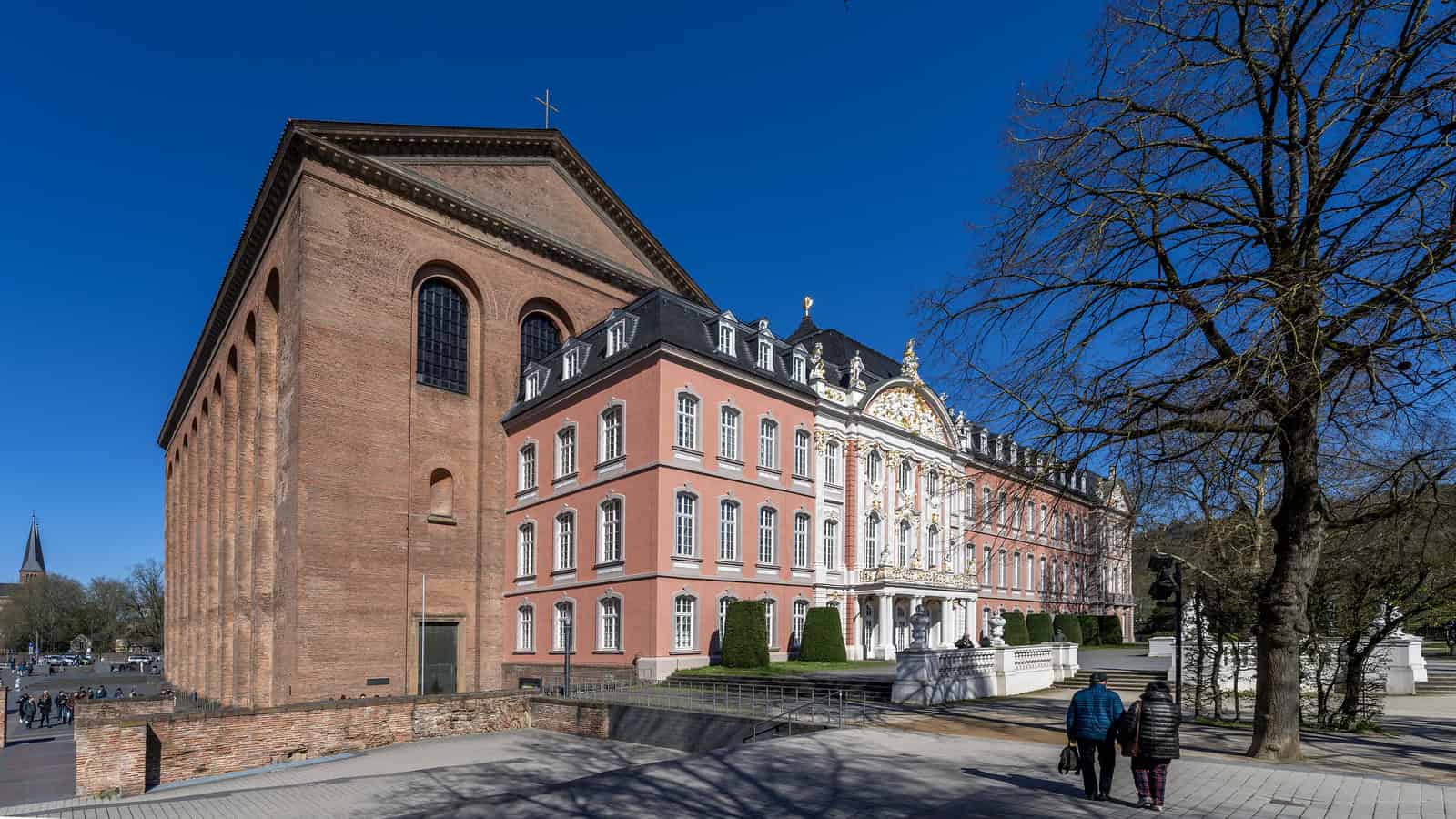 Stunning baroque palace architecture with intricate details and a historic brick church, set in a scenic location with leafless trees and visitors enjoying the outdoor space.