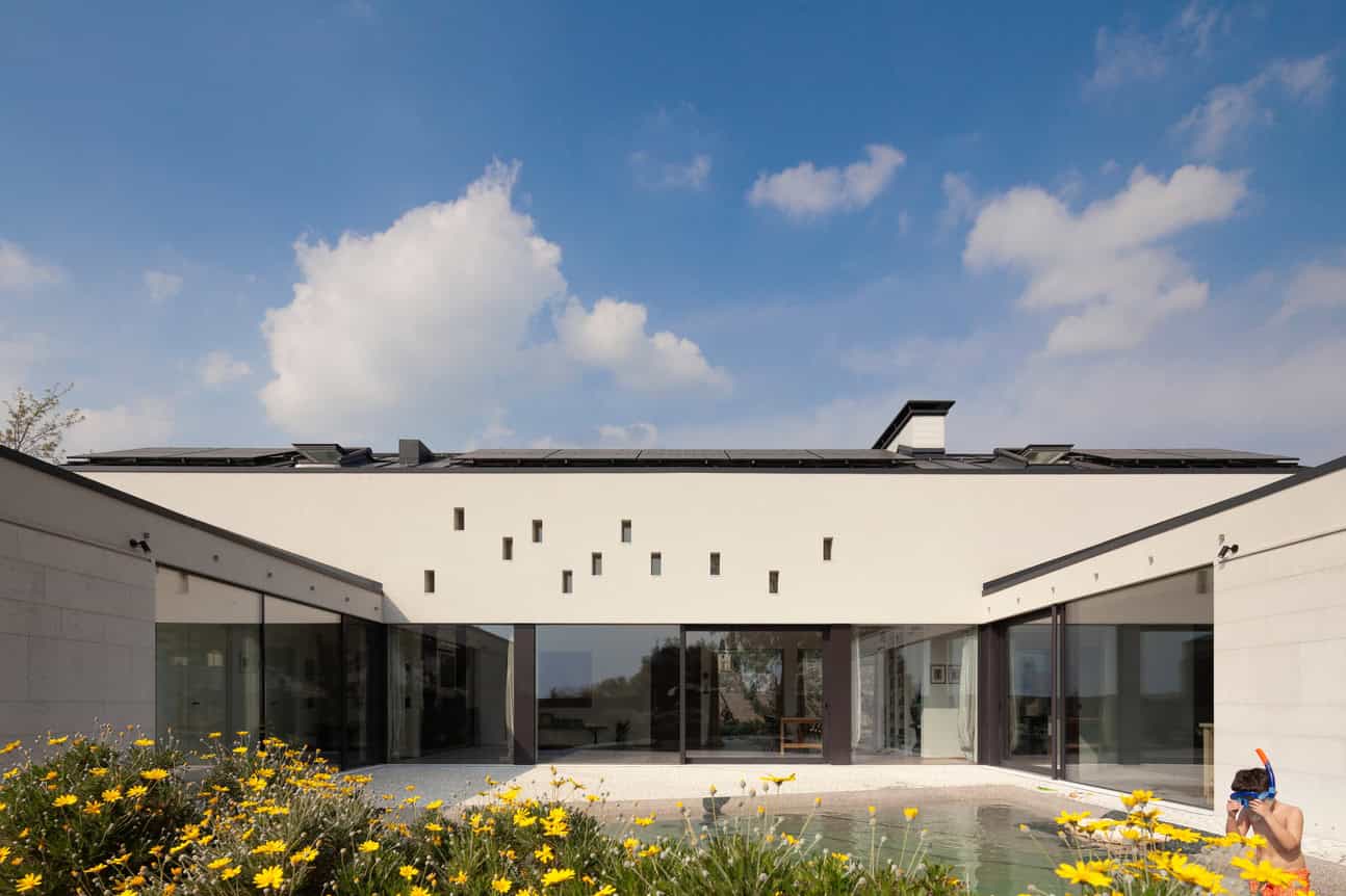 Modern minimalist house with clean lines, large glass windows, and contemporary architecture, set against a bright blue sky with landscaping including yellow flowers.