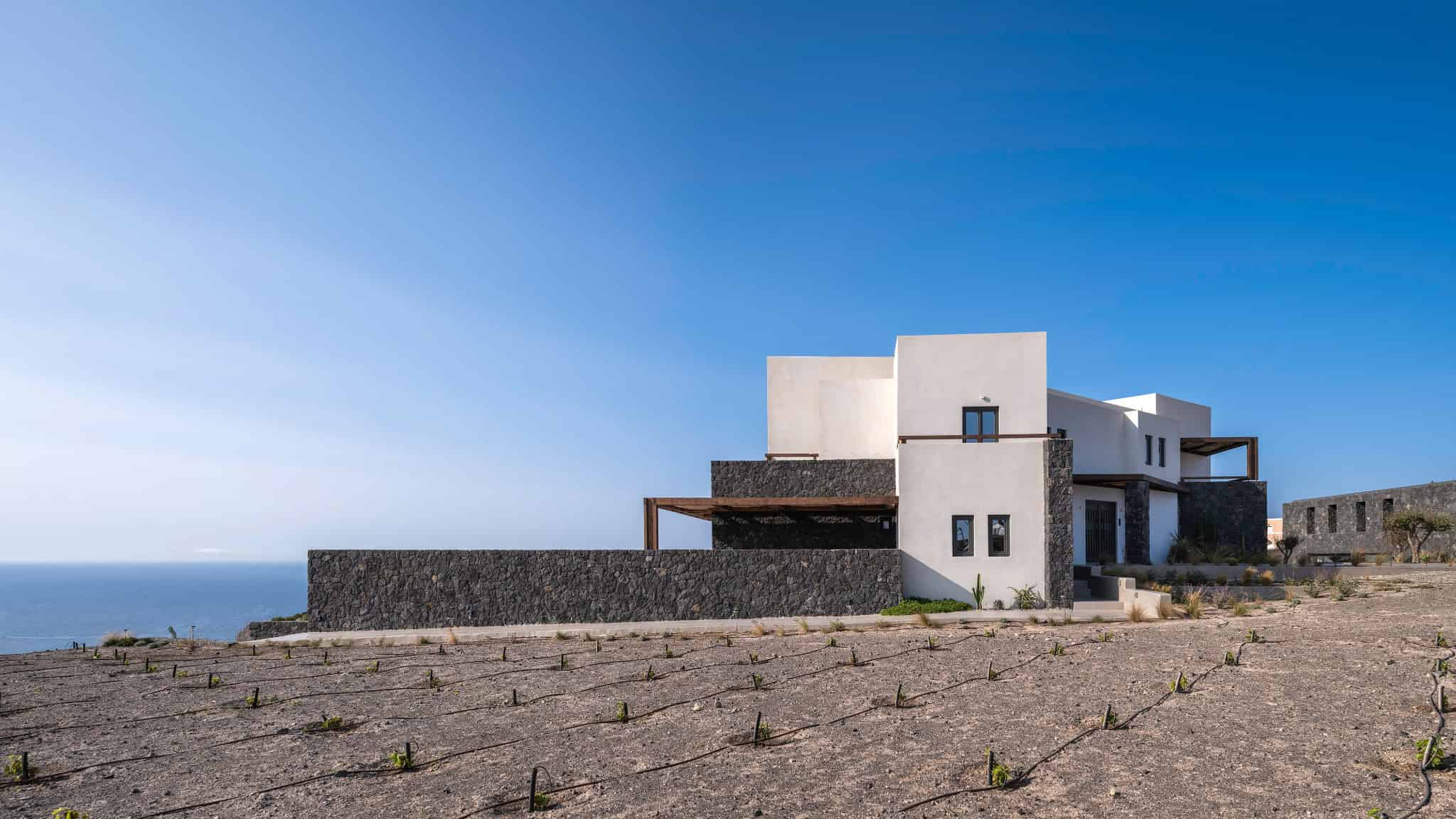 Modern minimalist house with white concrete walls and dark stone accents, overlooking the ocean, featuring clean lines and outdoor terraces, blending contemporary architecture with coastal landscape.