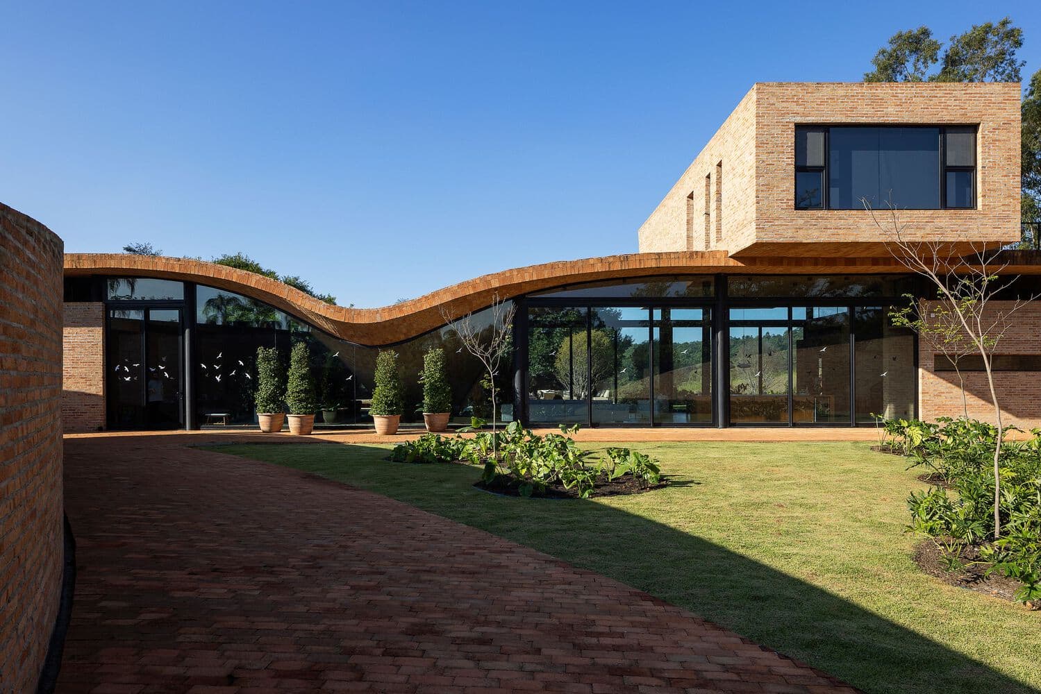 Contemporary modern house with curved brick roof and large glass windows, surrounded by a landscaped garden and paved walkway, showcasing innovative architecture and design.