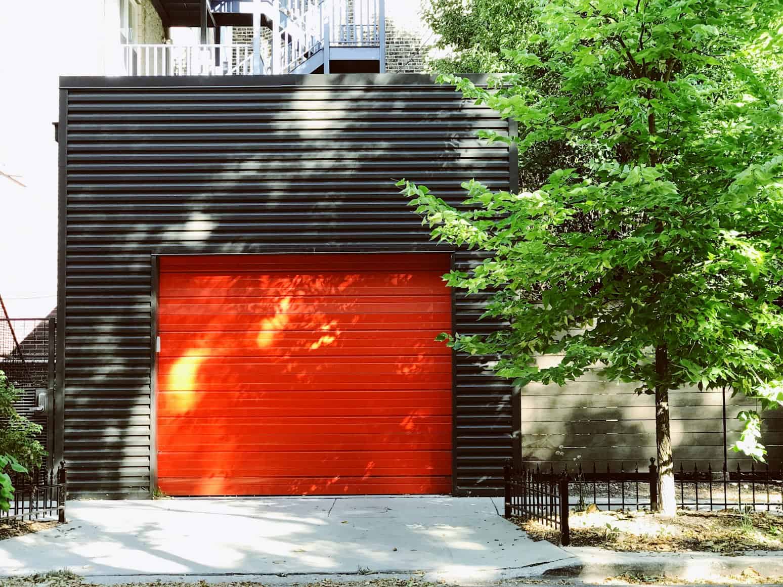 Modern residential garage with black ribbed metal siding and a bold red roll-up door, surrounded by lush green trees and a small black metal fence, exemplifying contemporary architecture and outdoor design.