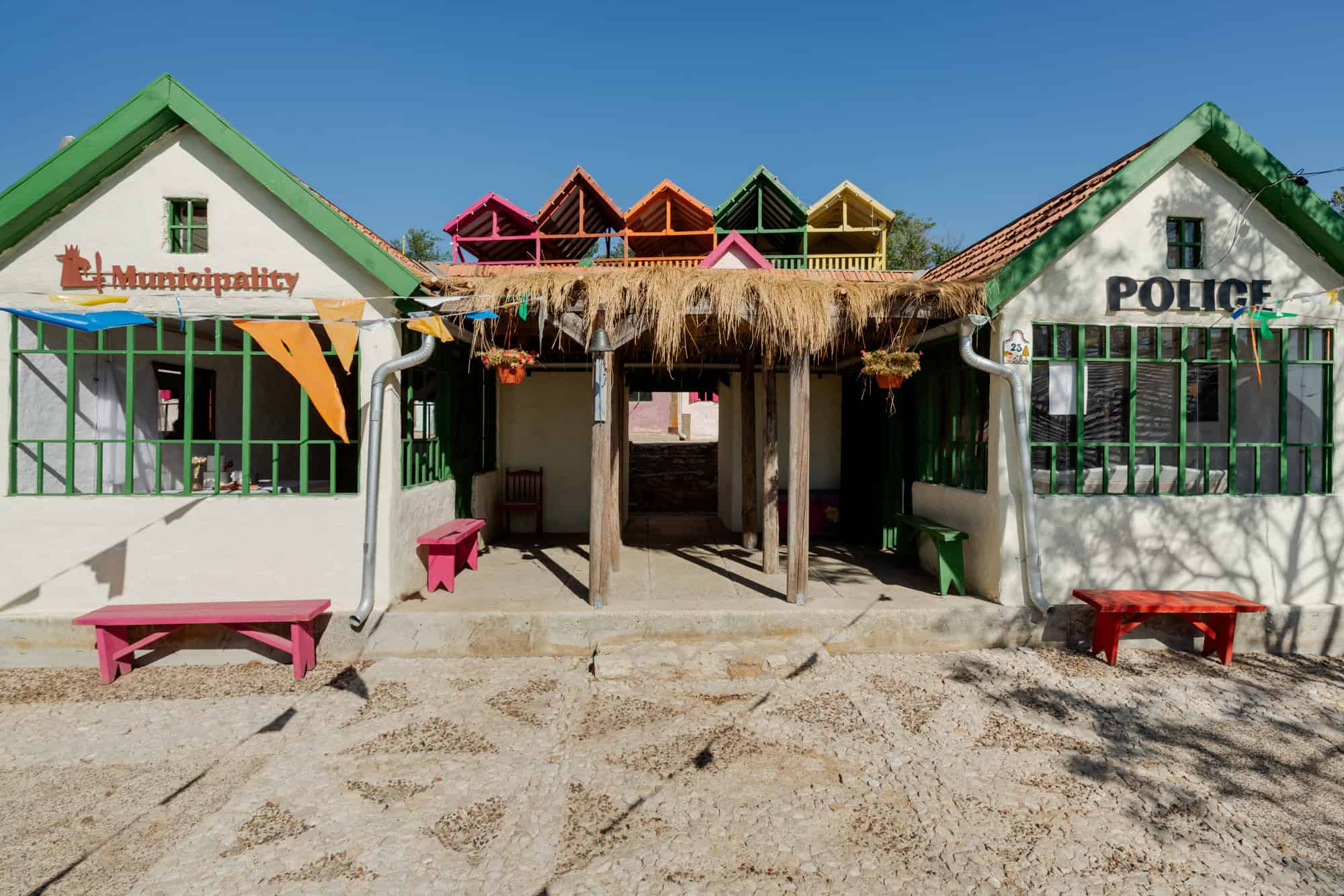 Colorful community building with vibrant painted benches, thatched roof, and bright architectural details in a rural setting, showcasing creative design in public spaces.