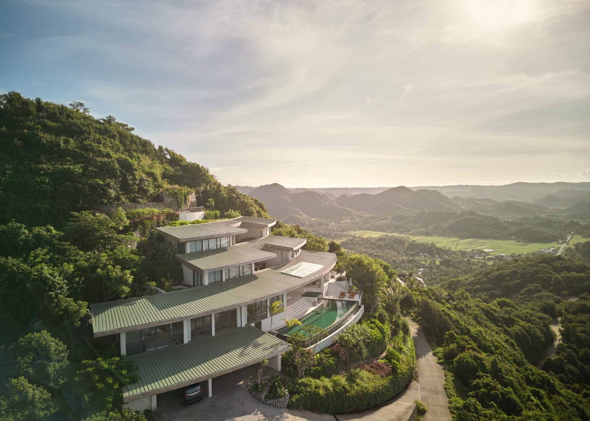Modern hillside luxury house with extensive glass windows and tiered design blending with lush green landscape, offering breathtaking mountain views and indoor-outdoor living.