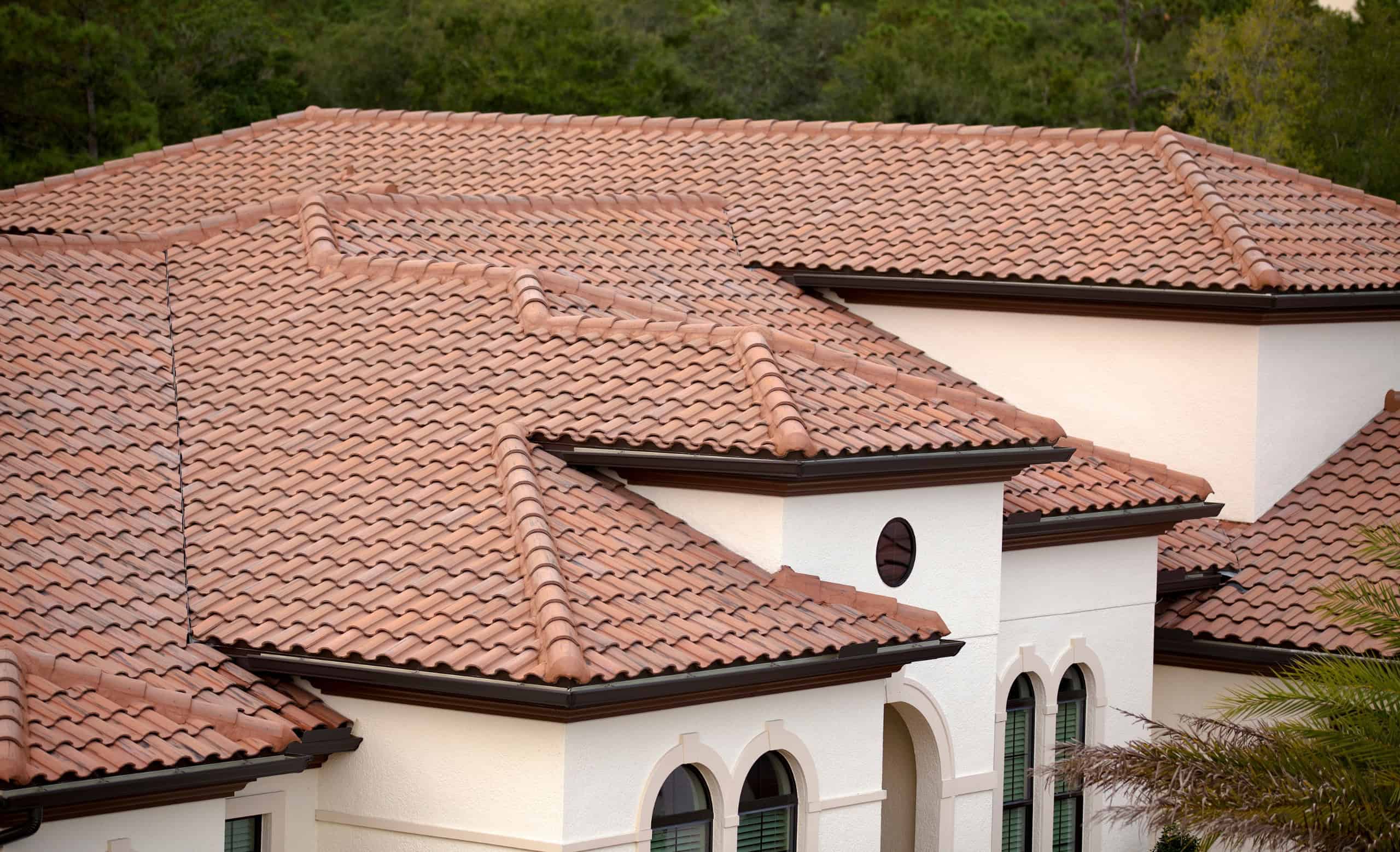Rich, terracotta clay tile roofing on a modern Mediterranean-style house with white stucco walls, surrounded by lush green trees, showcasing durable, stylish architecture and landscape integration.