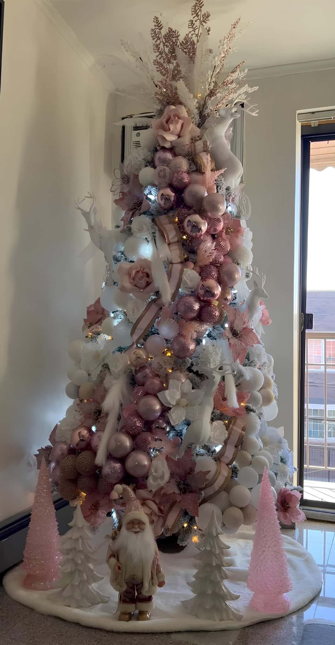 Pink and white Christmas tree decorated with flowers, ornaments, and fairy lights, featuring a Santa figurine at the base, in modern home interior with natural light from window.