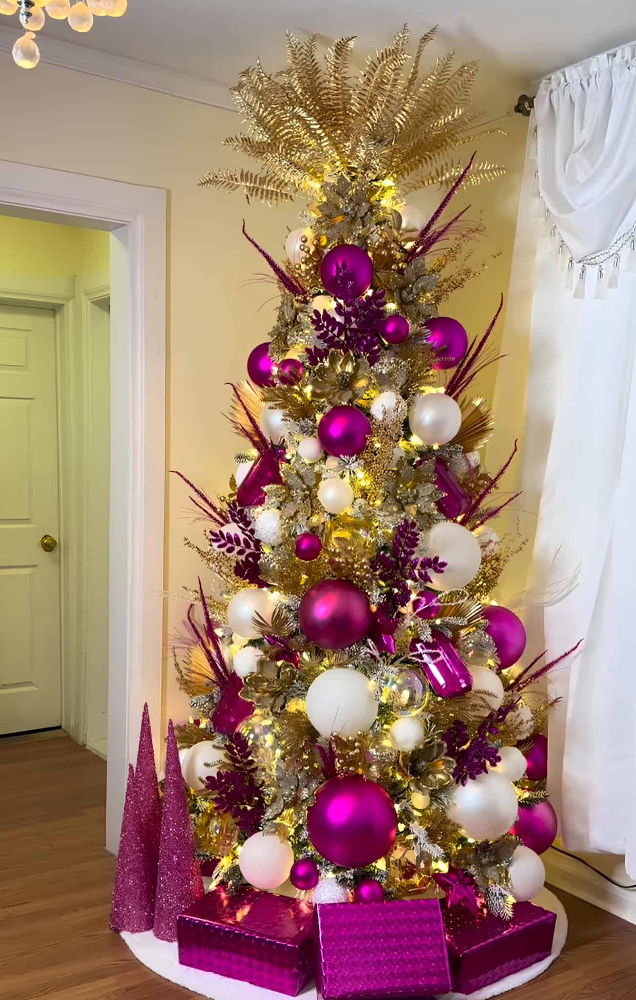 Lavender and gold Christmas tree decorated with purple and white ornaments, fairy lights, and gold foliage, placed indoors near white curtains, creating a festive holiday ambiance.