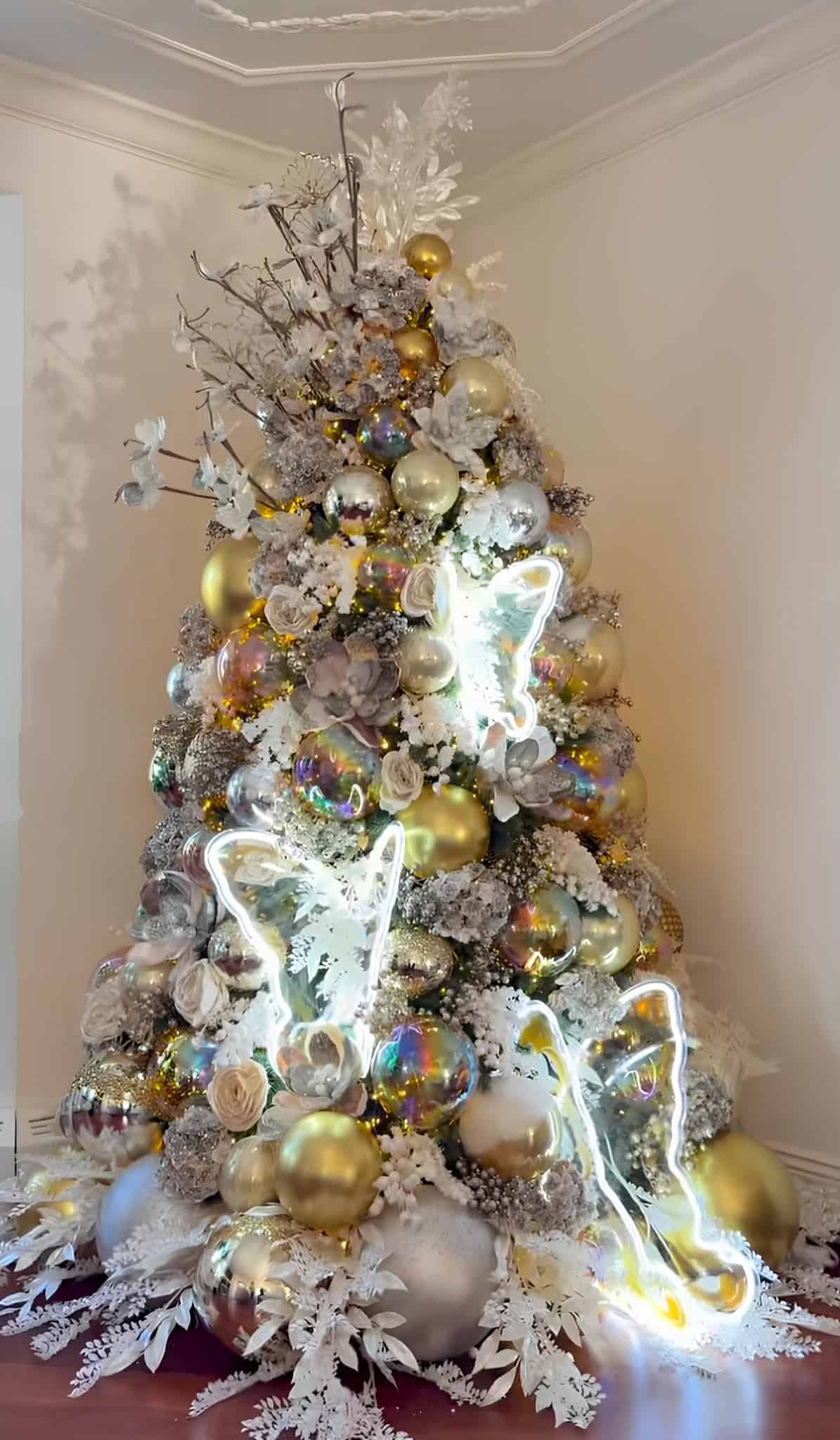 Ornamental Christmas tree decorated with gold and silver baubles, white floral accents, and LED lights, creating a luxurious holiday centerpiece, perfect for festive interior design.