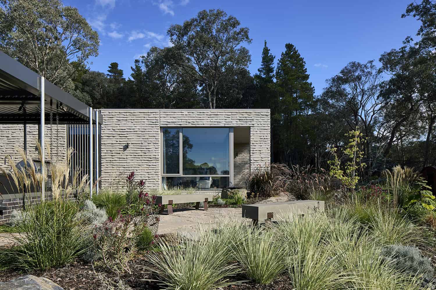 Contemporary modern house with minimalist architecture, flat roof, and white brick exterior, surrounded by desert landscaping with ornamental grasses and trees under a clear blue sky.