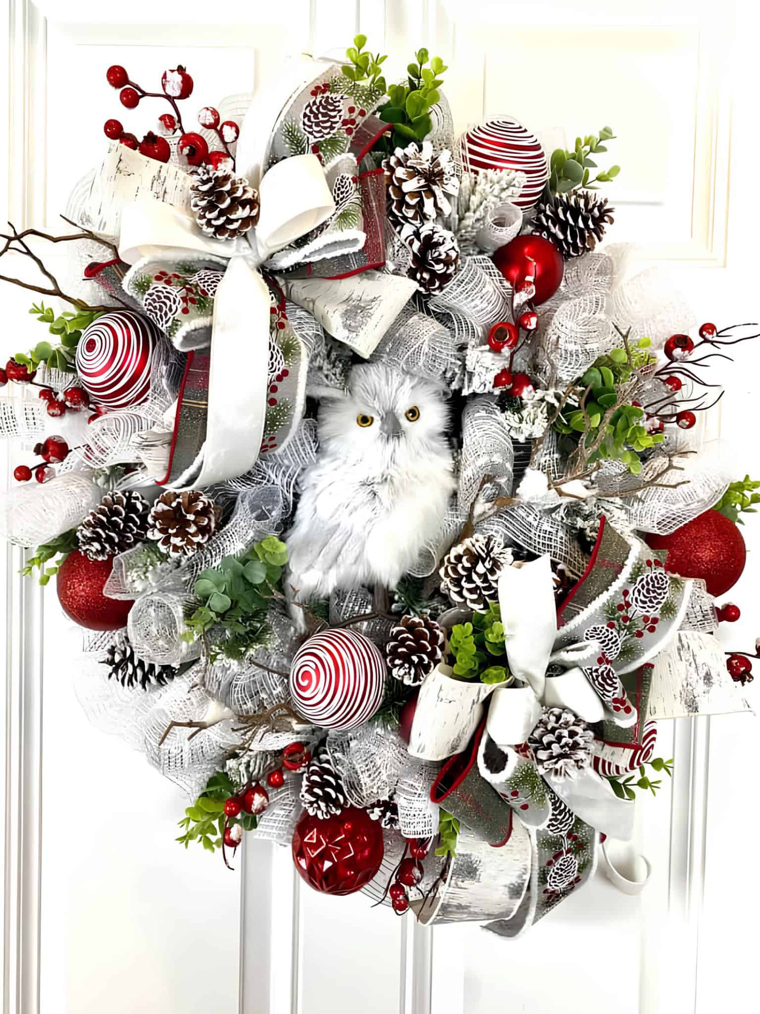White snowy owl nestled in a red and white ribbon Christmas wreath with pinecones and ornaments