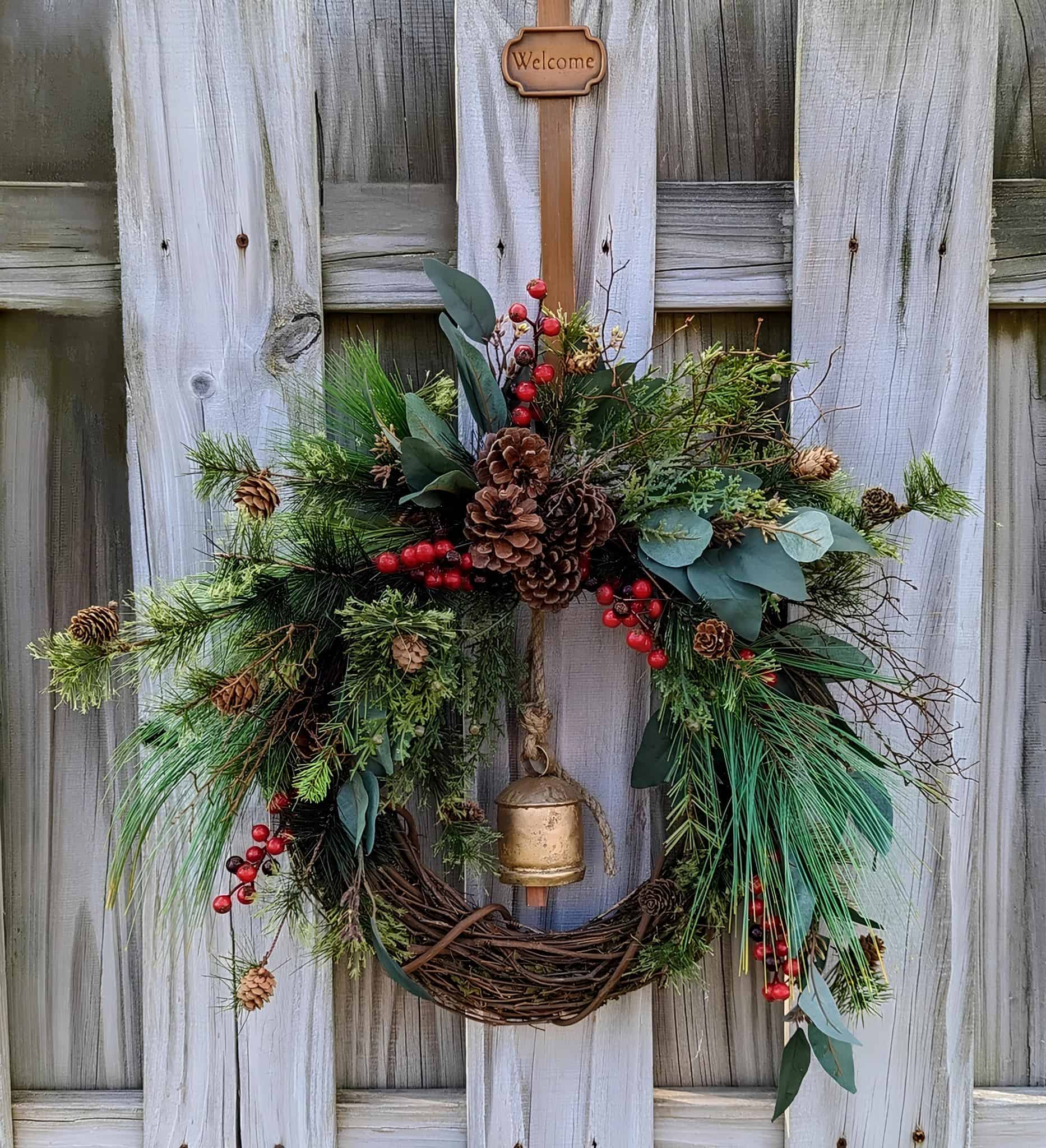 Vibrant holiday wreath with pinecones, red berries, green foliage, and a small bell, hanging on a weathered wooden fence for festive home decoration.