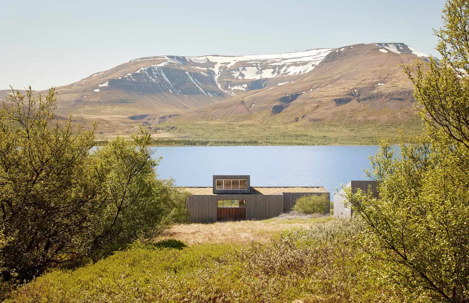 Modern lakeside house with minimalist design, surrounded by lush greenery and overlooking snow-capped mountains and a calm lake, exemplifying contemporary architecture in a natural setting.