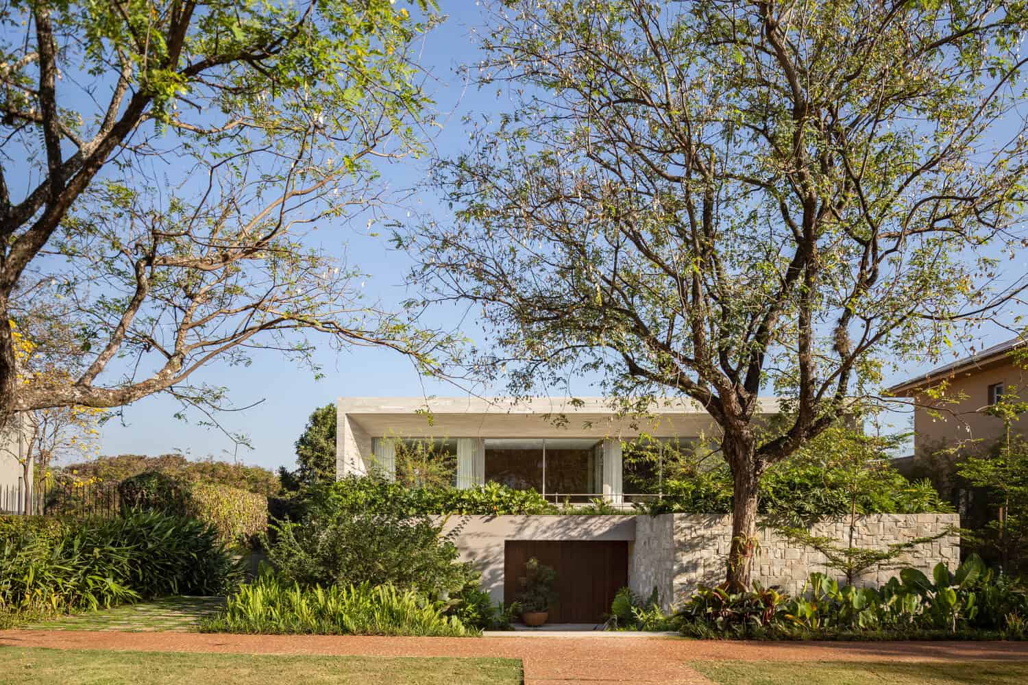 Modern minimalist house with stone accents and large glass windows nestled among lush greenery and mature trees, showcasing contemporary architecture with clean lines and natural materials.