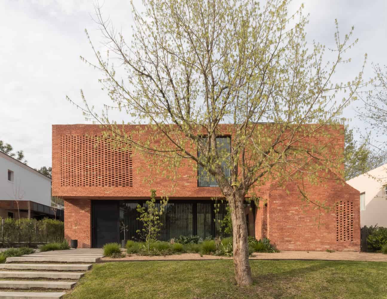 Contemporary red brick house with geometric design and large glass windows, surrounded by greenery and landscaped yard. Modern architecture emphasizing clean lines and natural materials.