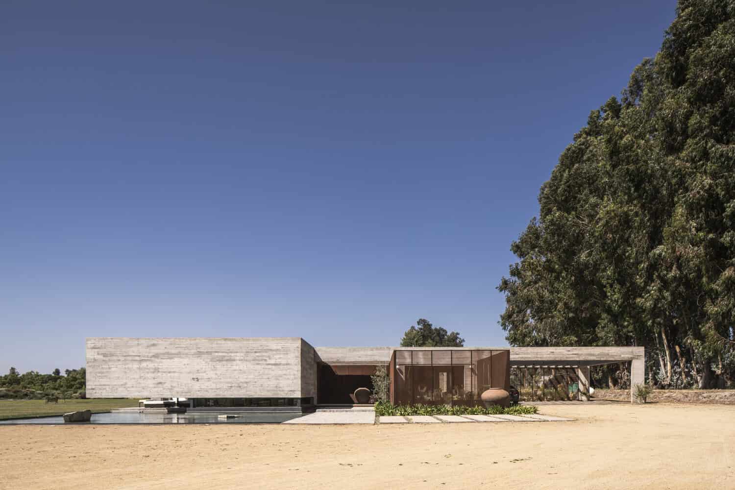 Contemporary minimalist house with modern architectural design, concrete and rust-colored metal accents, large glass windows, situated in a natural landscape with trees and open space.