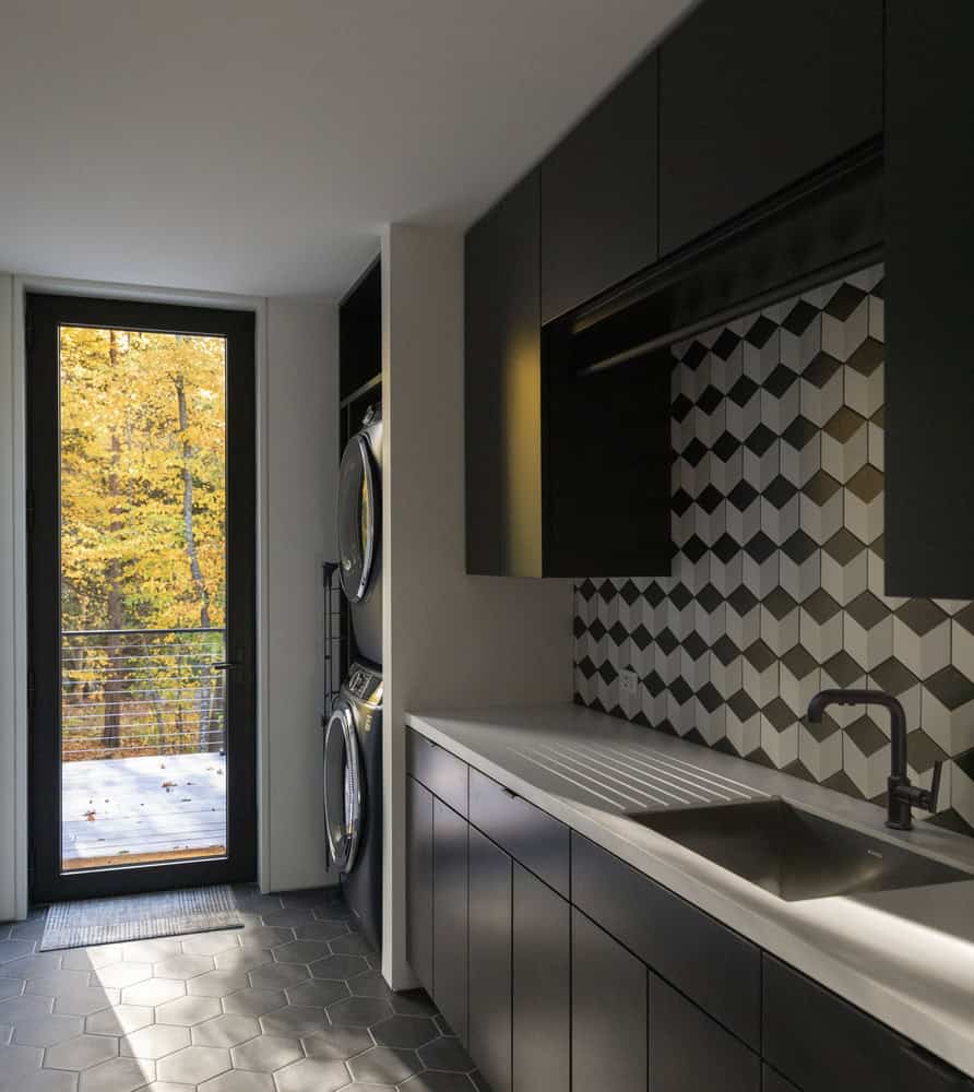 Sleek modern laundry room with black cabinetry and geometric backsplash design, featuring front-loading washer and dryer, black faucet, and natural light from glass door with outdoor autumn trees in background.