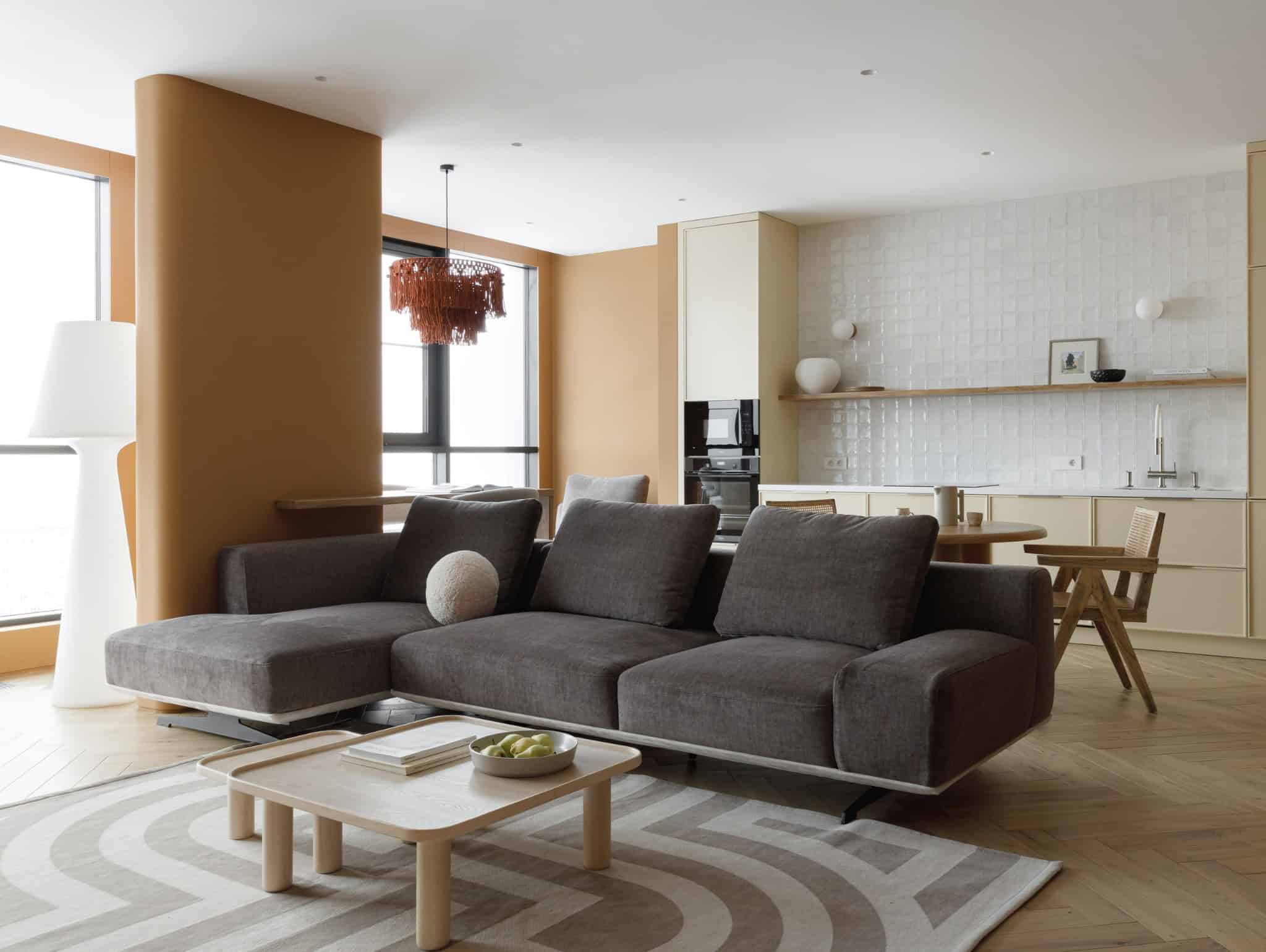 Contemporary open-plan living room with modern kitchen, beige walls, dark gray sofa, and natural wood accents, featuring stylish decor, large windows, and minimalist design elements.