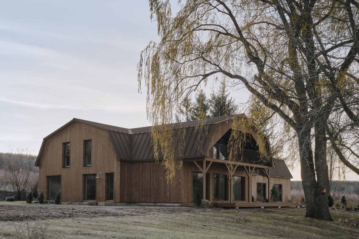 Maison Melba by Atelier L’Abri with covered barn porch and large tree in foreground in Frelighsburg, Quebec