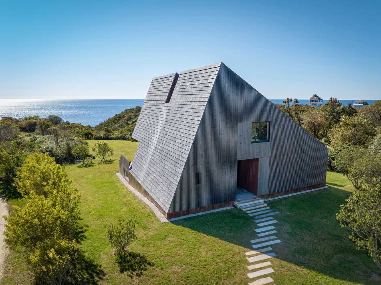 Modern wooden A-frame house with sloped roof near the coast, surrounded by greenery and ocean view, showcasing contemporary architectural design and sustainable building practices.