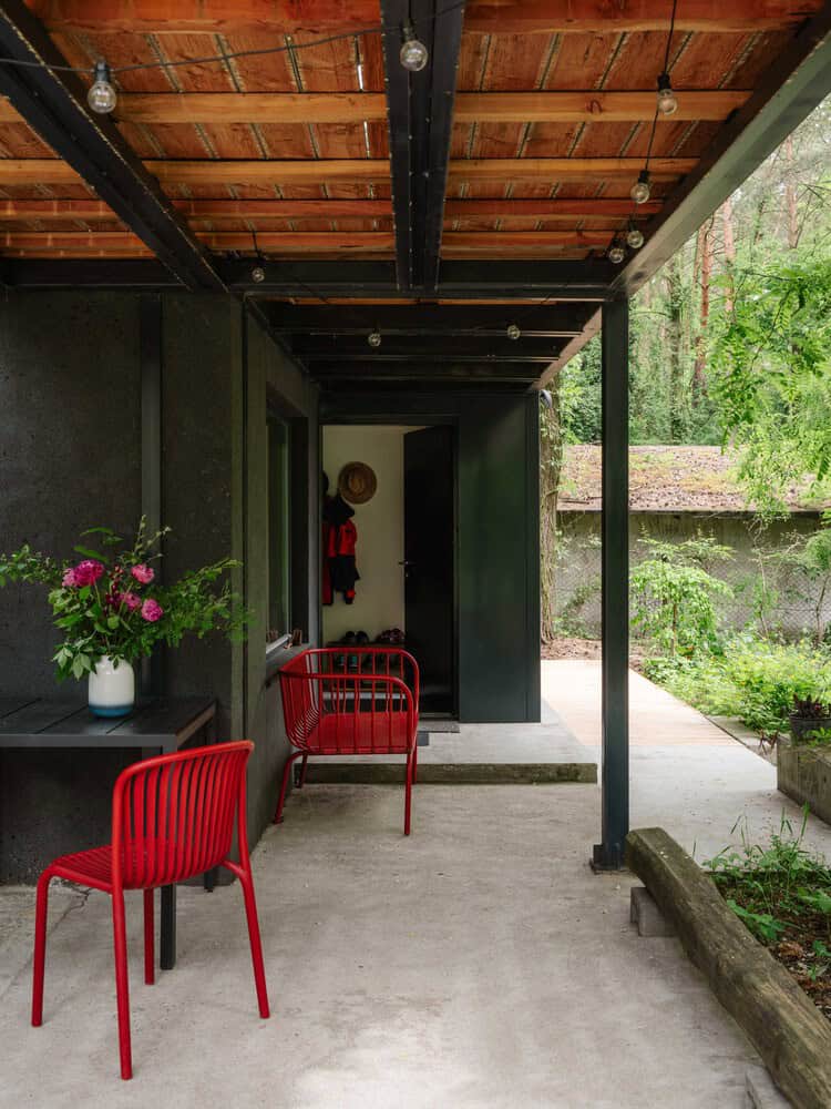 Modern outdoor patio with sleek black walls, wooden ceiling, stylish red chairs, and lush greenery, exemplifying contemporary architecture and innovative design in outdoor living spaces.