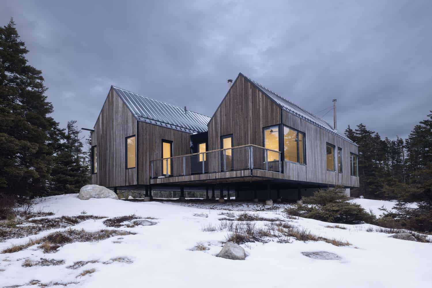 Evening view of Tide Bound House by Peter Braithwaite Studio featuring warm interior light, timber cladding, and raised deck in Indian Harbour, Nova Scotia, Canada
