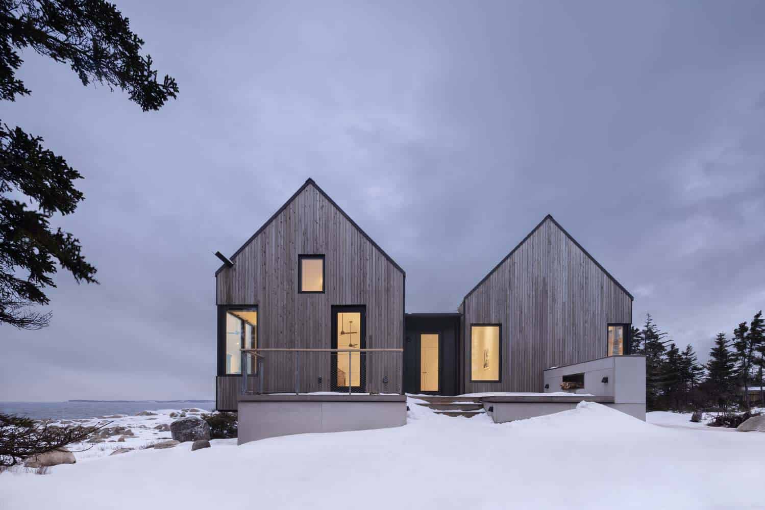 Front facade of Tide Bound House by Peter Braithwaite Studio with three gabled volumes glowing at dusk in Indian Harbour, Nova Scotia