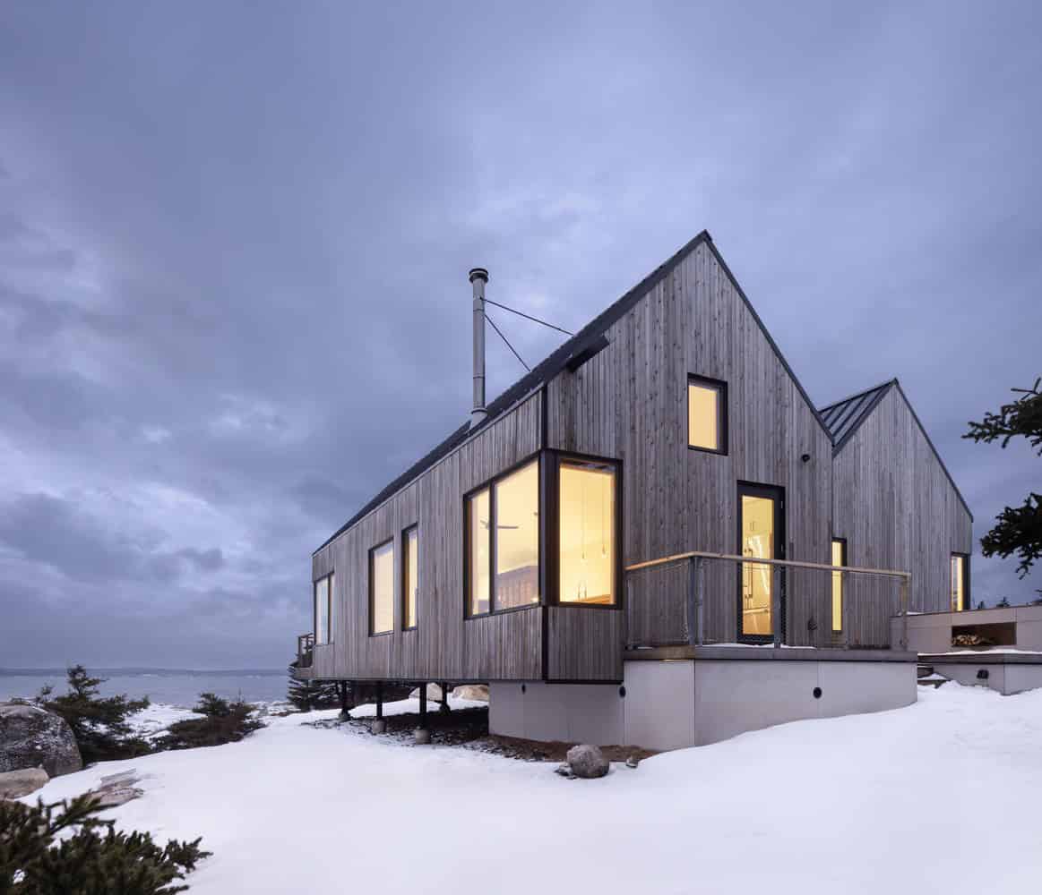 Corner view of Tide Bound House by Peter Braithwaite Studio showing illuminated gable ends and large window openings at dusk in Indian Harbour, Nova Scotia
