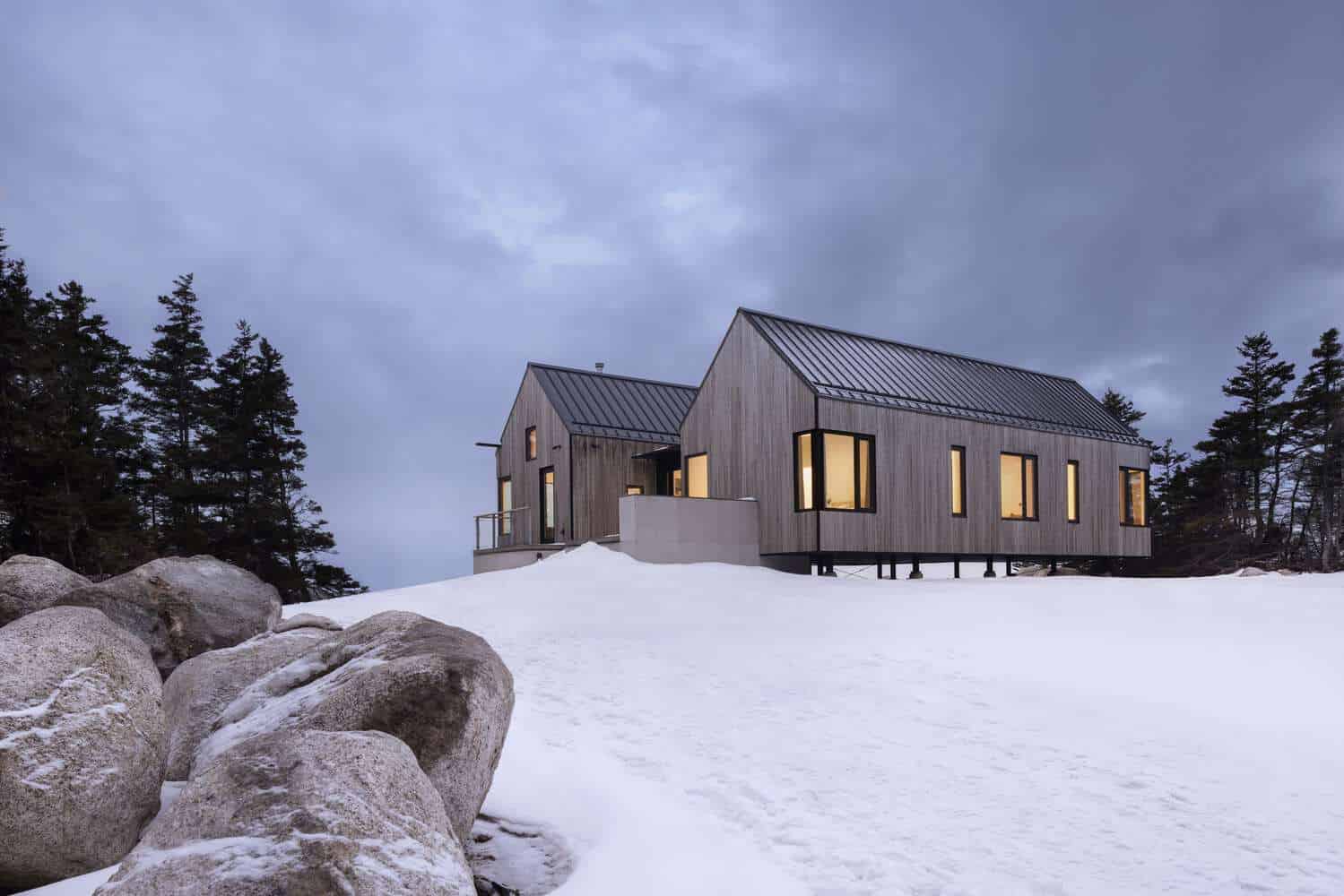 Tide Bound House by Peter Braithwaite Studio resting above a snowy rocky slope at dusk in Indian Harbour, Nova Scotia