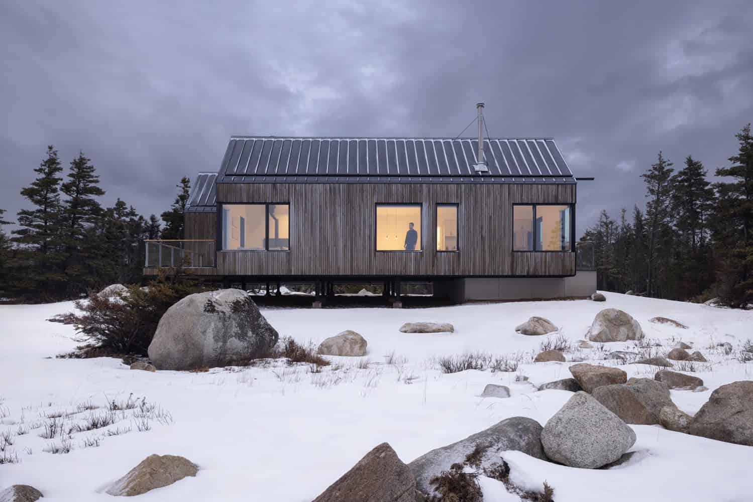 Side elevation of Tide Bound House by Peter Braithwaite Studio illuminated from within, standing on a raised structure over snowy terrain in Indian Harbour, Nova Scotia
