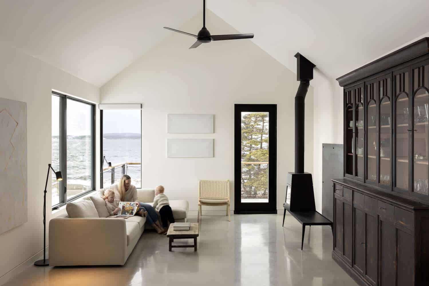 Family living area inside Tide Bound House by Peter Braithwaite Studio with sofa, picture windows and ocean views in Indian Harbour