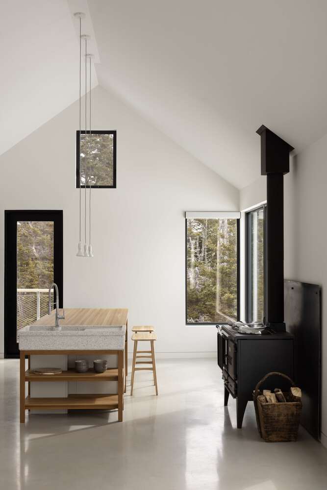 Minimal interior kitchen of Tide Bound House by Peter Braithwaite Studio featuring wood stove, island and forest views in Indian Harbour, Nova Scotia