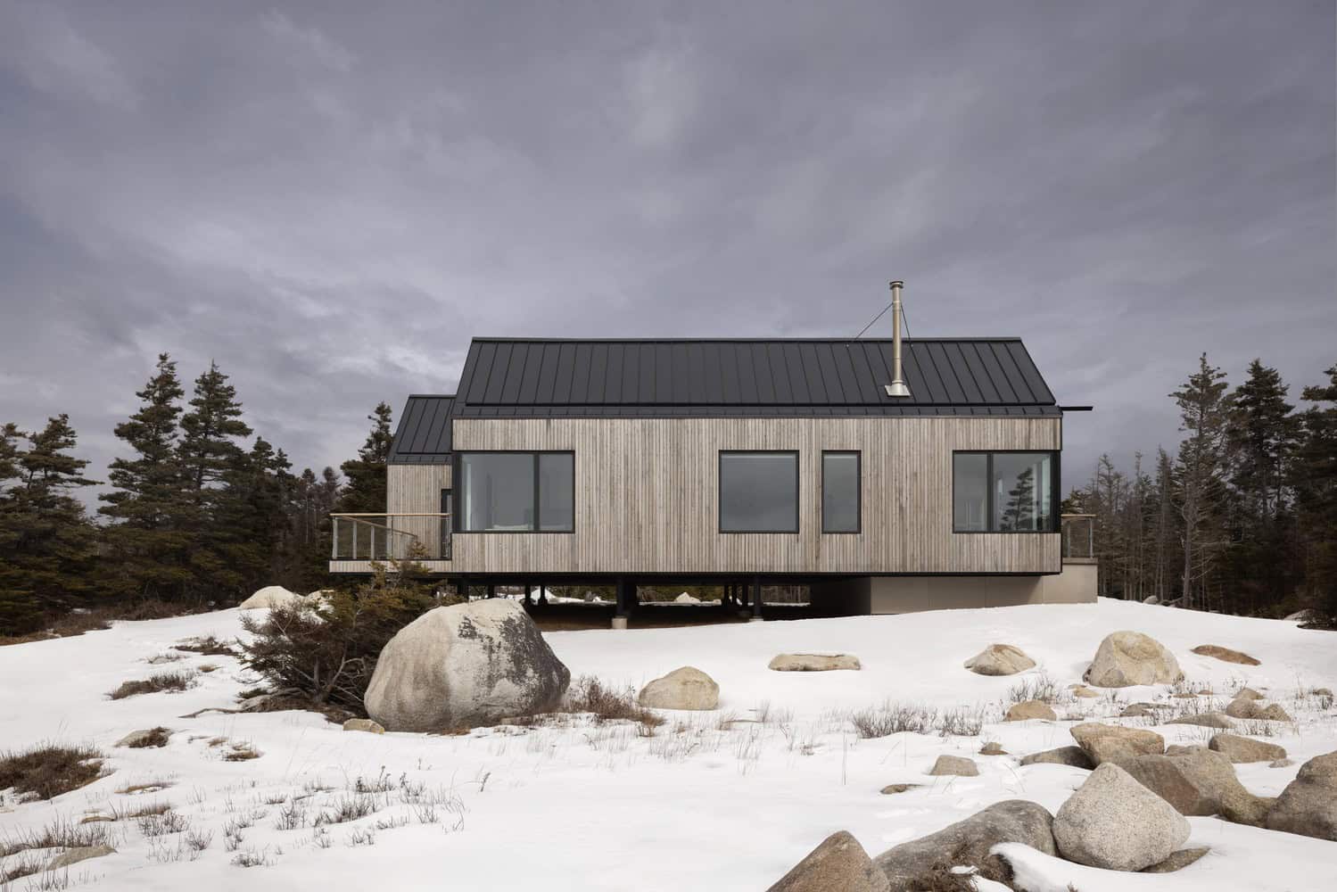 Side elevation of Tide Bound House by Peter Braithwaite Studio on a raised foundation above the snowy coastal terrain in Indian Harbour, Nova Scotia, Canada