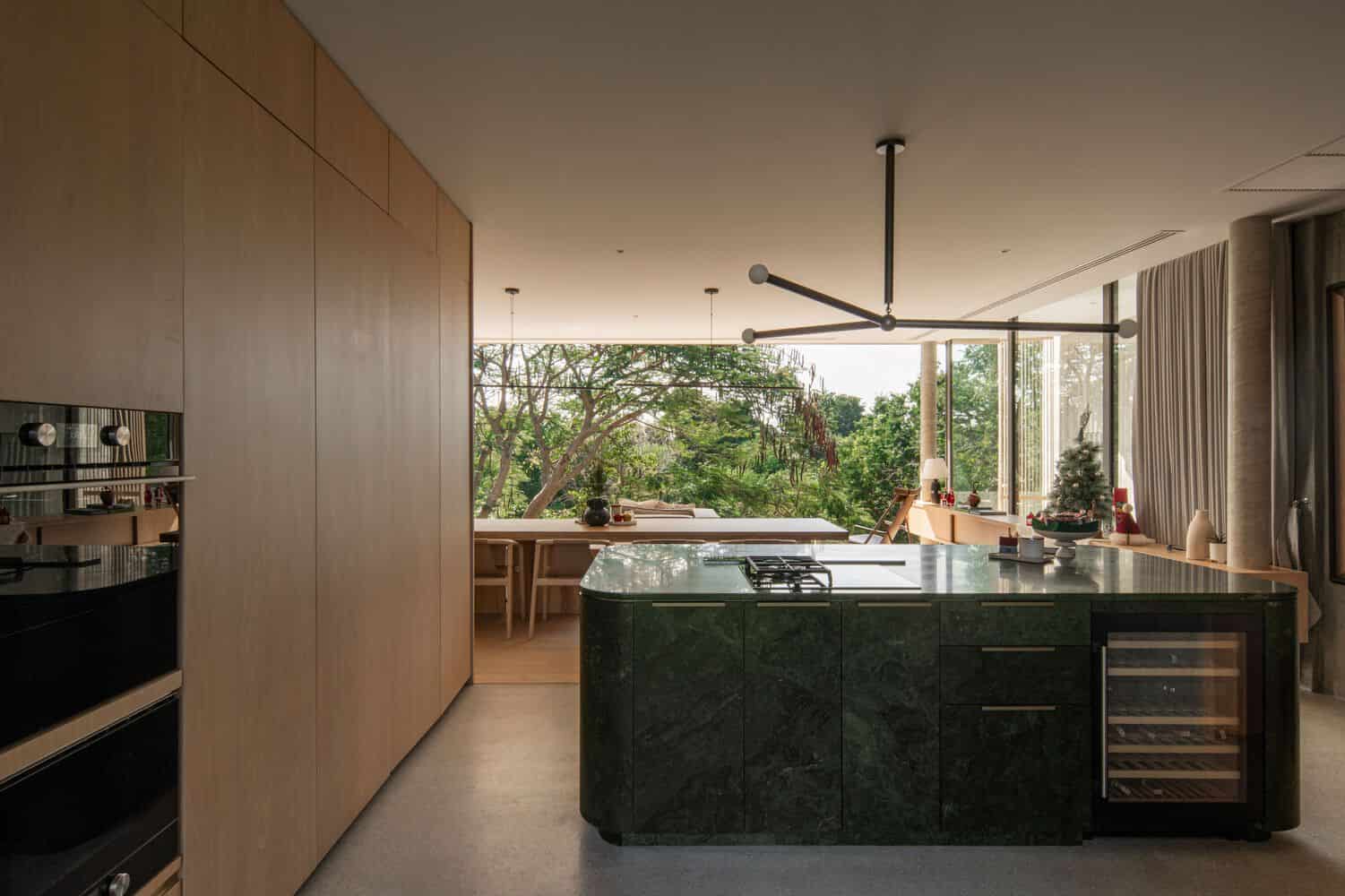 Modern open-concept kitchen with natural wood cabinets, green marble island, and large windows overlooking lush outdoor greenery, perfect for contemporary architecture and stylish interior design.